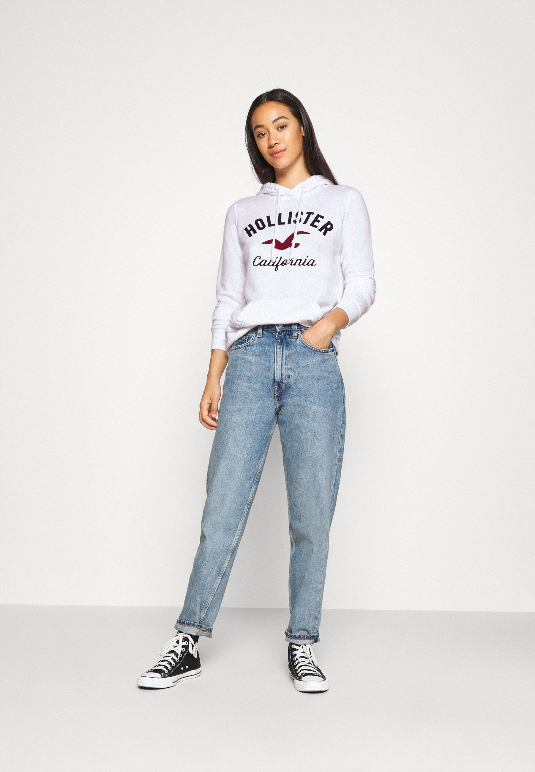 黑紅配色Hollister標誌 壁紙 — 一位年輕女性穿著白色Hollister連帽衫、藍色牛仔褲和黑色運動鞋，在純白色工作室背景前自信地擺姿勢。