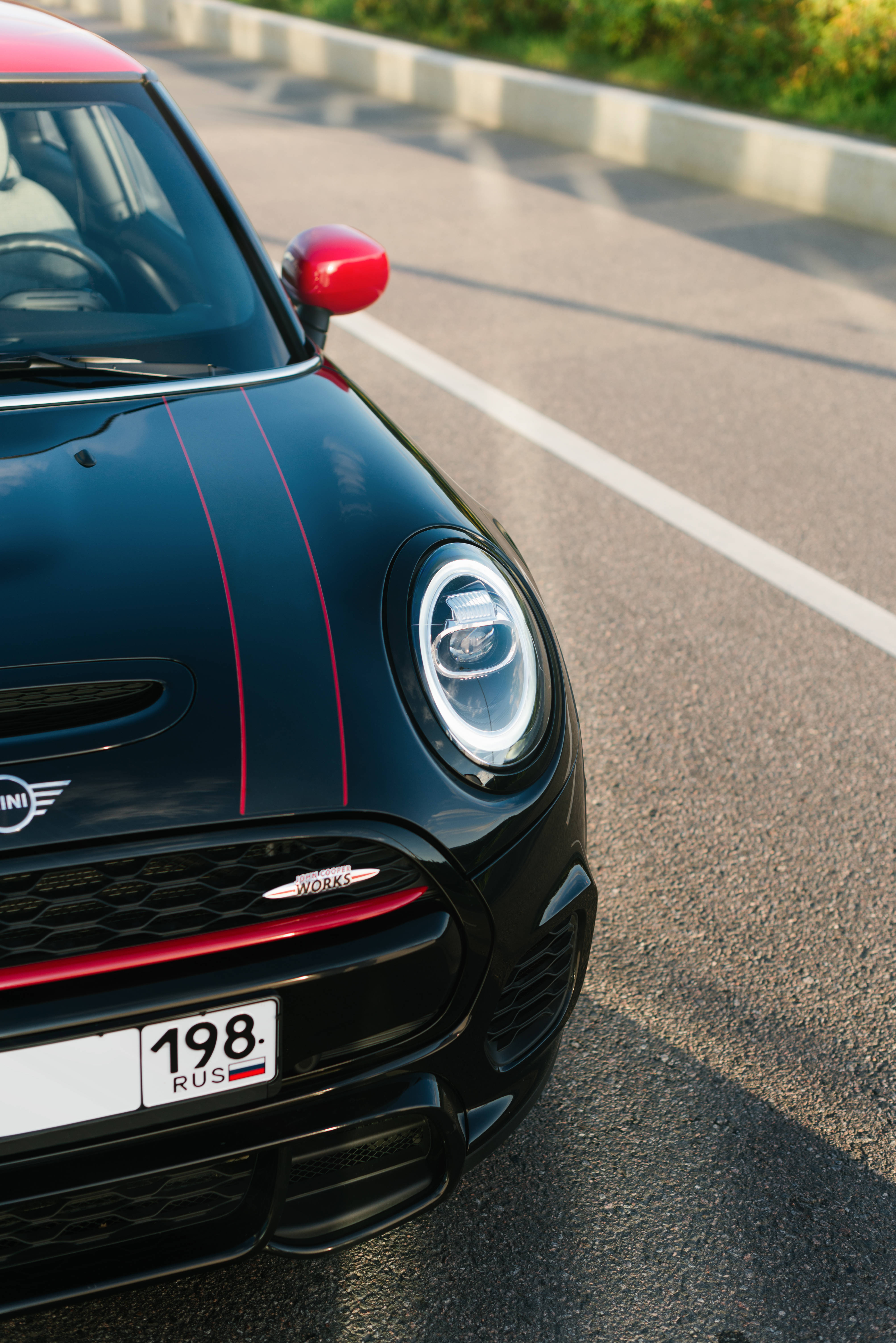 黑紅配色Mini Cooper車身上的Mini Cooper標誌 壁紙 — 一輛時尚的黑色MINI Cooper帶有紅色點綴，停泊在陽光普照的瀝青路上，背景有綠化植物。