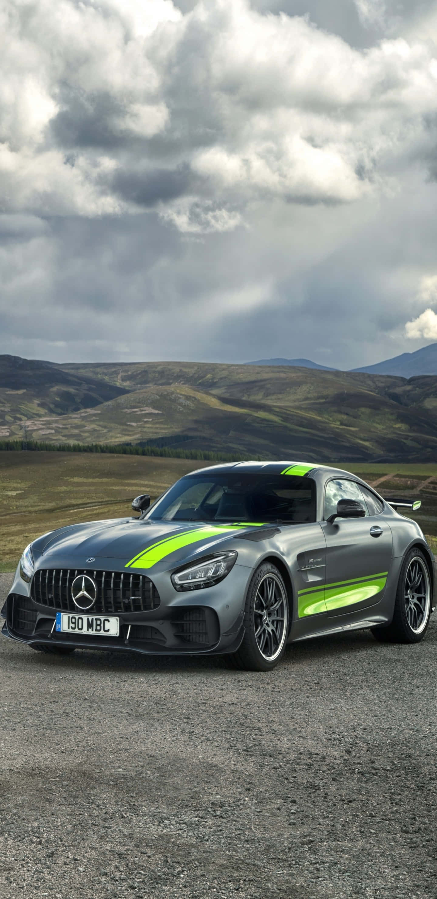 黑綠配色梅賽德斯-奔馳 Pixel 3XL AMG GT-R 背景 壁紙 — 一輛時尚的灰色Mercedes-AMG GT R，帶有螢光綠色條紋，停在碎石上，背景是戲劇性的多雲天空和起伏的山丘。