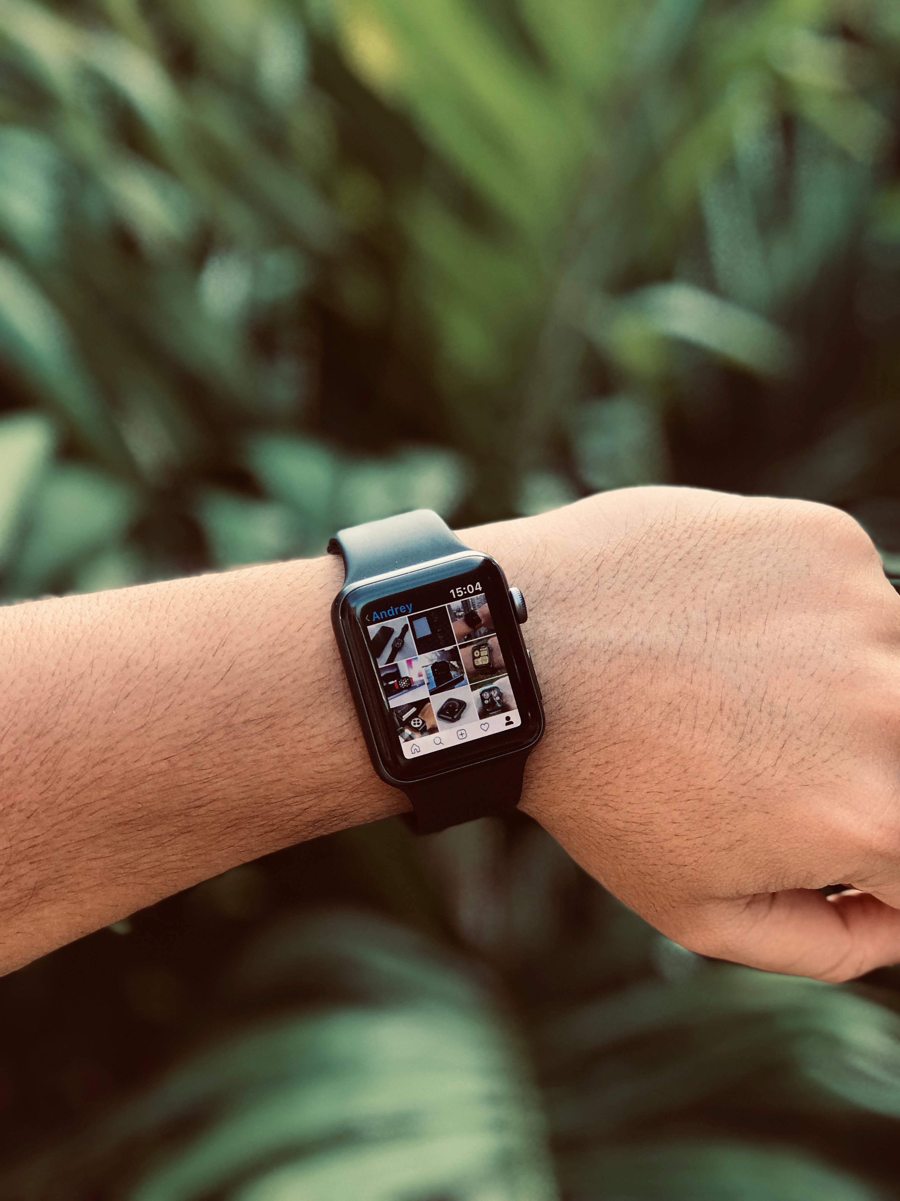 黑色 Apple 智能手錶 壁紙 — 一個人手腕的特寫，戴著智能手錶，在模糊的綠色樹葉背景前顯示相片庫。
