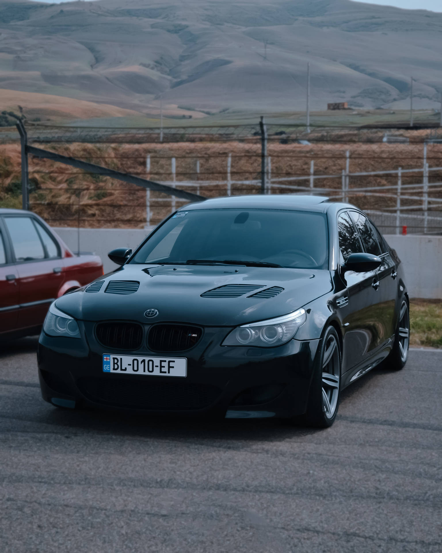黑色BMW M5停在圍欄附近的瀝青路上，背景有丘陵。