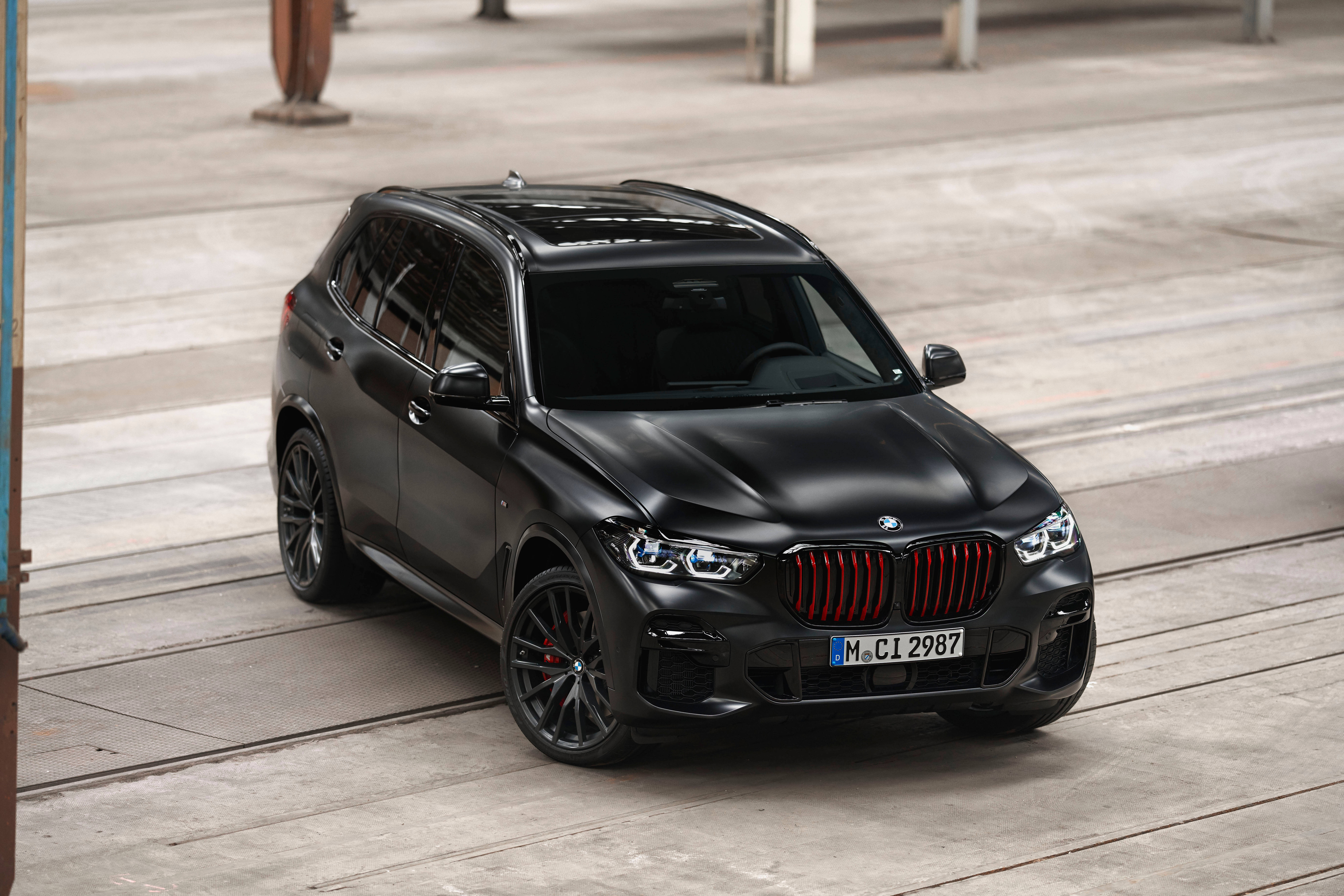 黑色 BMW X6 前視圖 壁紙 — 一輛啞光黑色BMW X5 M Competition SUV，帶紅色飾件，停泊在工業橫樑下的混凝土上。
