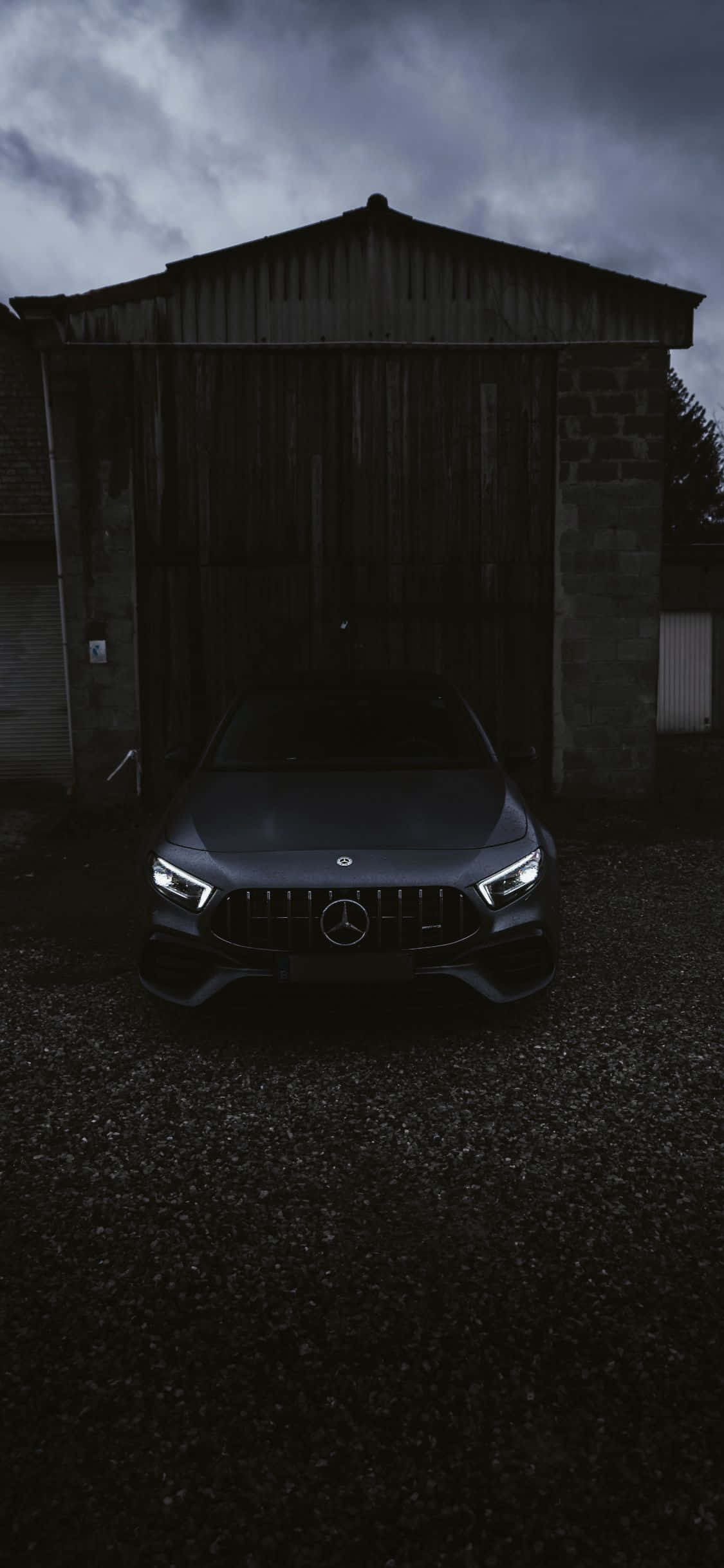 黑色 iPhone Xs Mercedes AMG 前視背景 壁紙 — 一架時尚的深灰色 Mercedes-AMG 跑車在風雨交加的天空下，於一間風化的穀倉前，在車頭燈下閃耀。