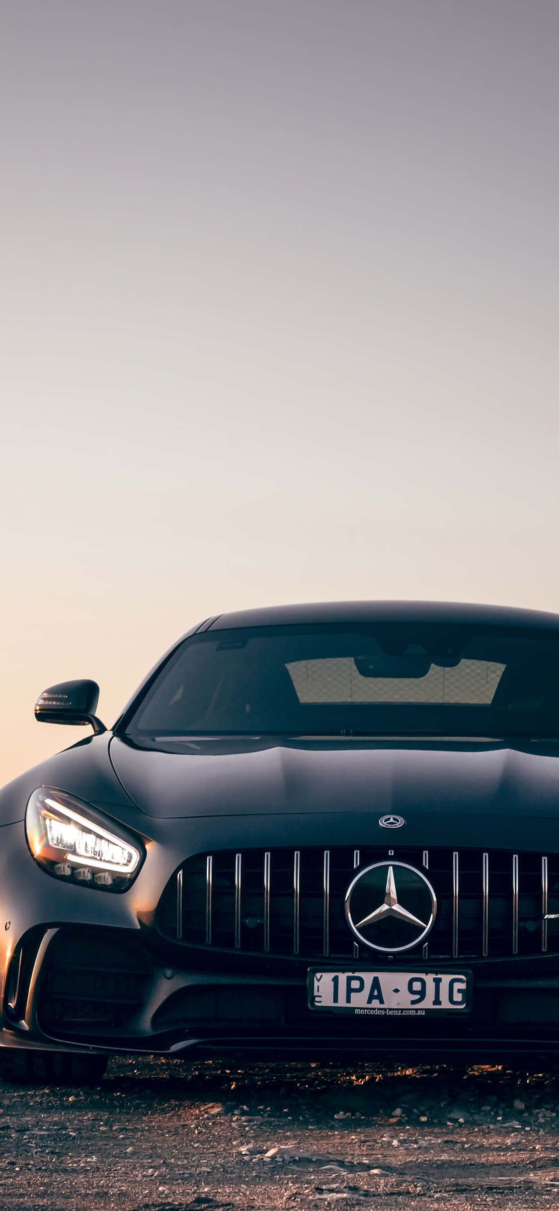 黑色 iPhone Xs Mercedes AMG 背景 壁紙 — 一輛時尚的黑色Mercedes-AMG GT停在碎石上，在柔和暮色天空下，車頭燈閃閃發光。