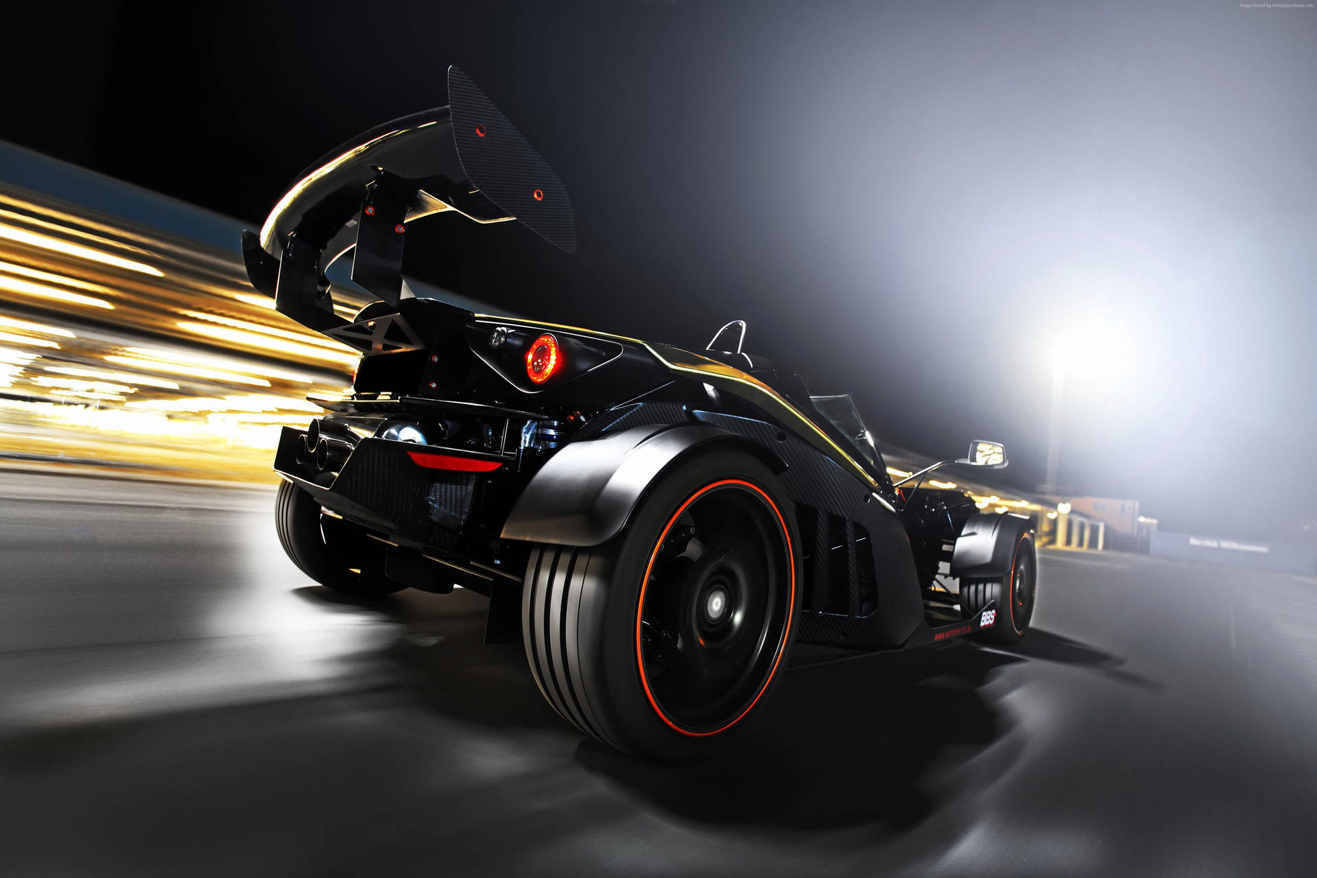 黑色 KTM X-Bow 賽車 壁紙 — 一輛黑色和金色的跑車帶有發光的橙色點綴，在夜間賽道上疾馳，留下模糊的光跡。