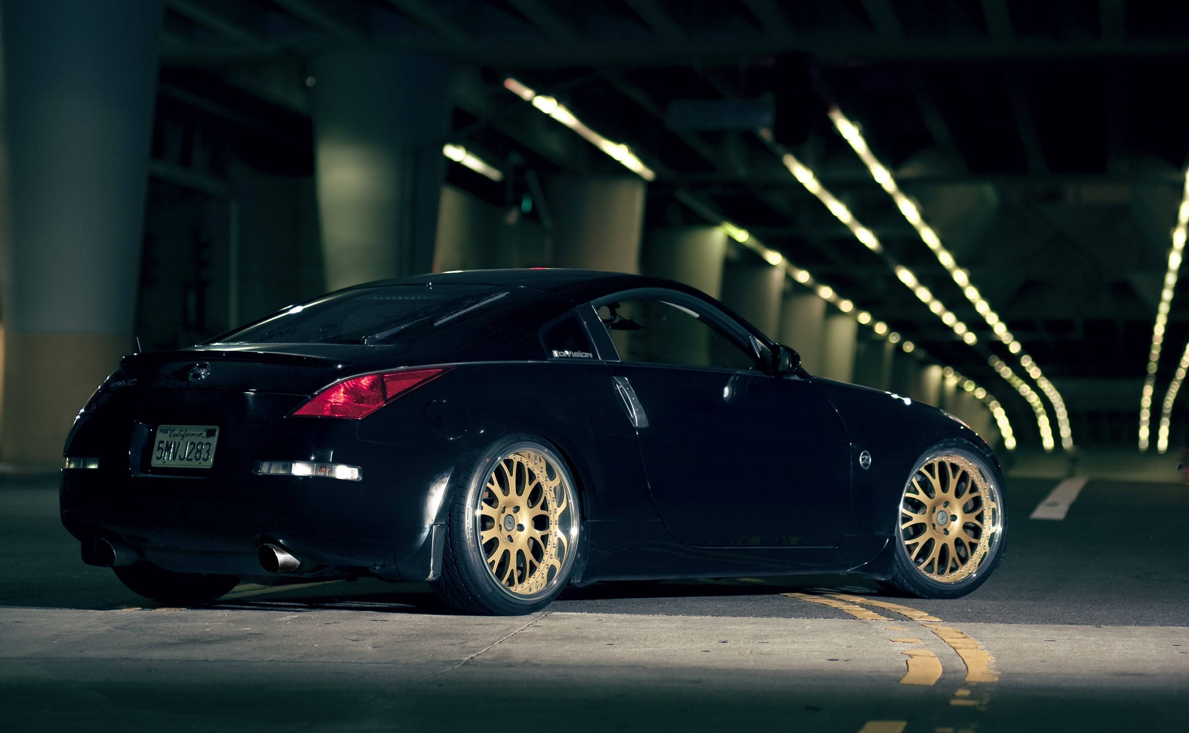黑色 Nissan 350z iPhone 鎖屏 壁紙 — 一輛時尚的黑色跑車配金色輪圈，停放在燈光昏暗、掛滿發光燈串的隧道下。