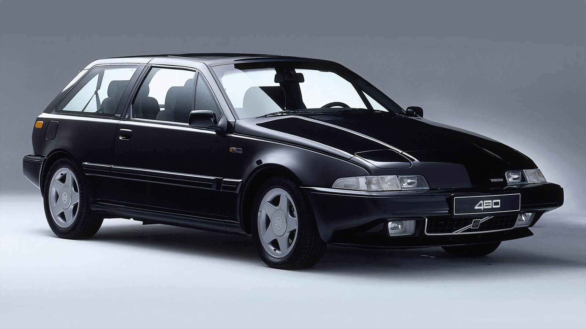 黑色480系列Volvo iPhone相片 壁紙 — 一輛時尚的黑色富豪480掀背車在攝影棚燈光下，於簡約灰色背景前閃閃發光。
