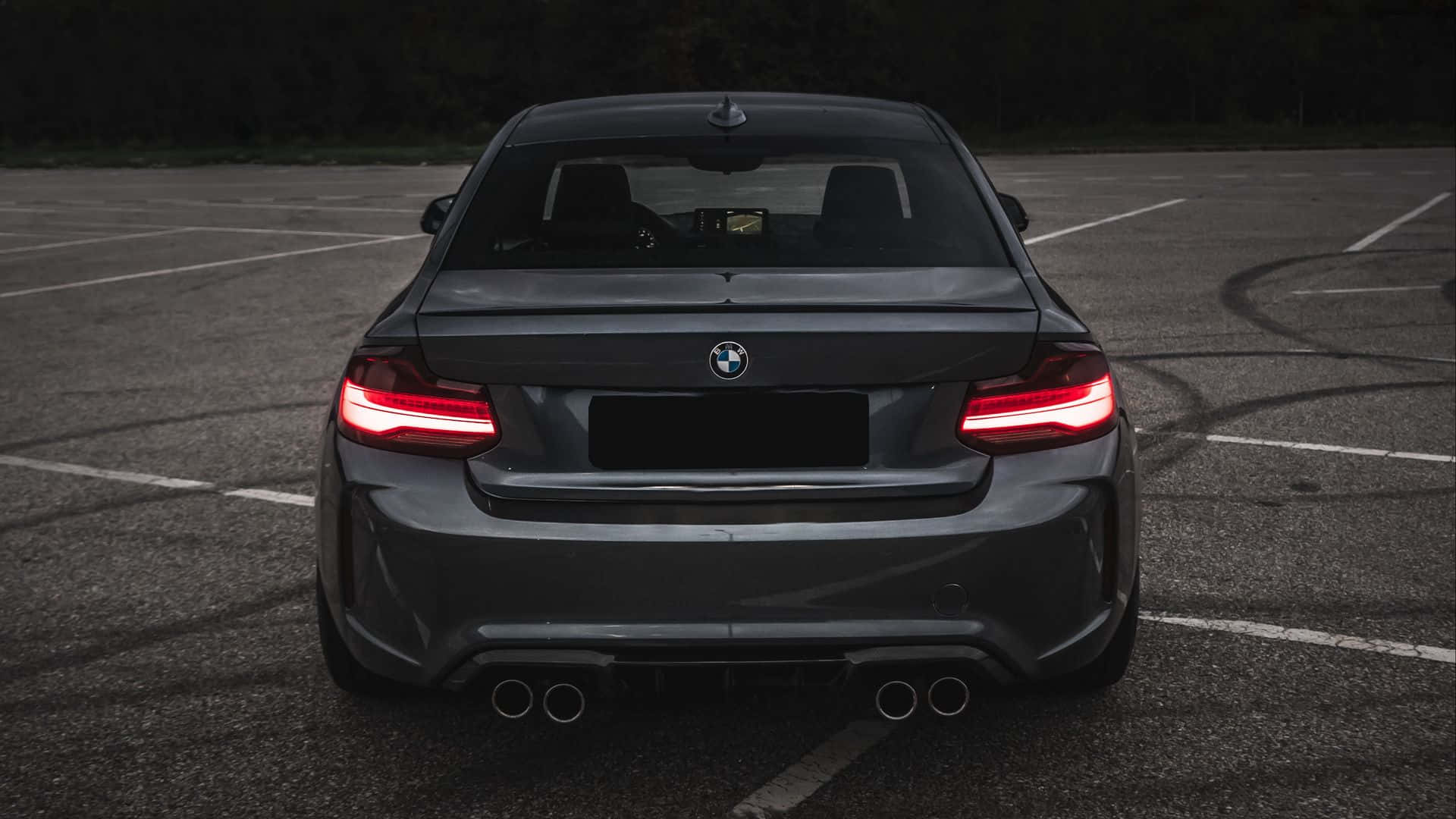 黑色BMW M3後視圖 1920x1080 M系列背景 壁紙 — 一架時尚的深灰色BMW M2跑車，帶有發光的紅色尾燈，停泊在黃昏時空無一人的瀝青停車場。