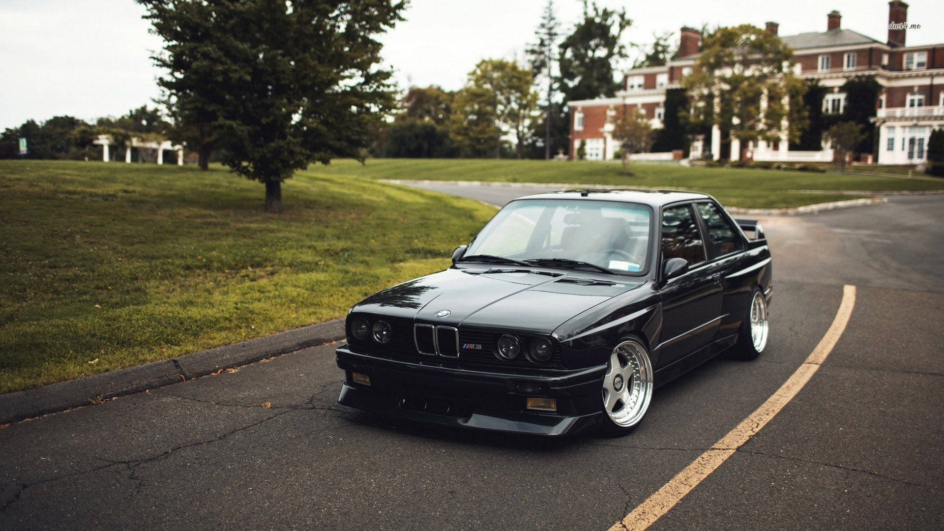 黑色BMW M3行駛於住宅區道路 壁紙 — 一輛時尚的黑色BMW E30 M3跑車停泊在彎曲的瀝青路上，旁邊是修剪整齊的綠色草坪和宏偉的磚砌房屋。