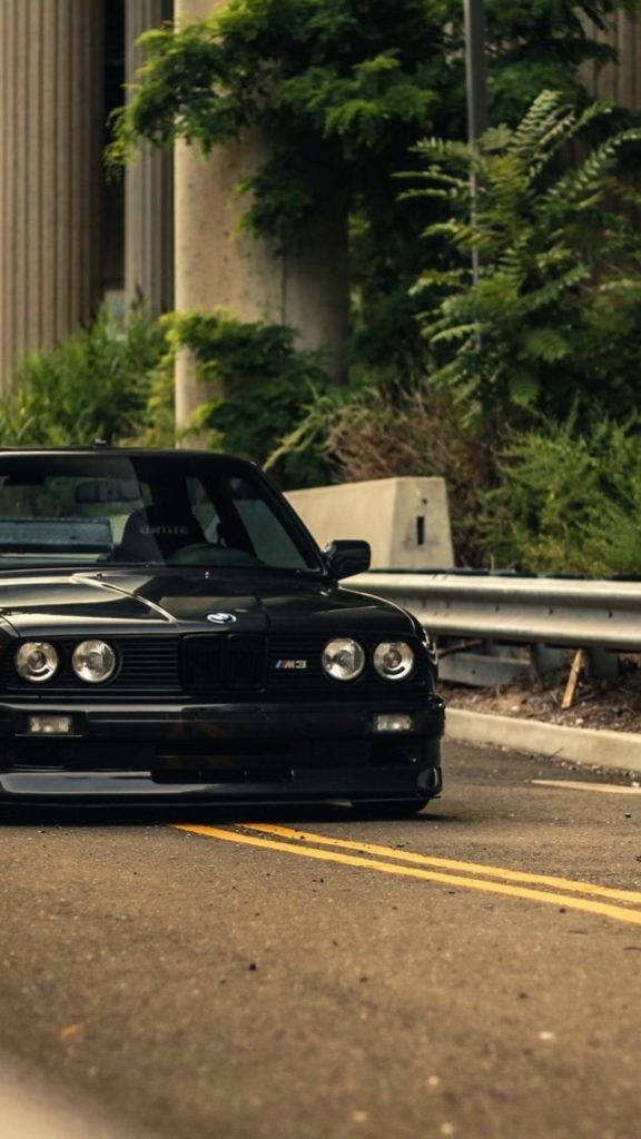 黑色BMW M3駛過風景公路 壁紙 — 一架時尚黑色BMW M3跑車停泊在路邊，背景有茂密綠植和混凝土柱。