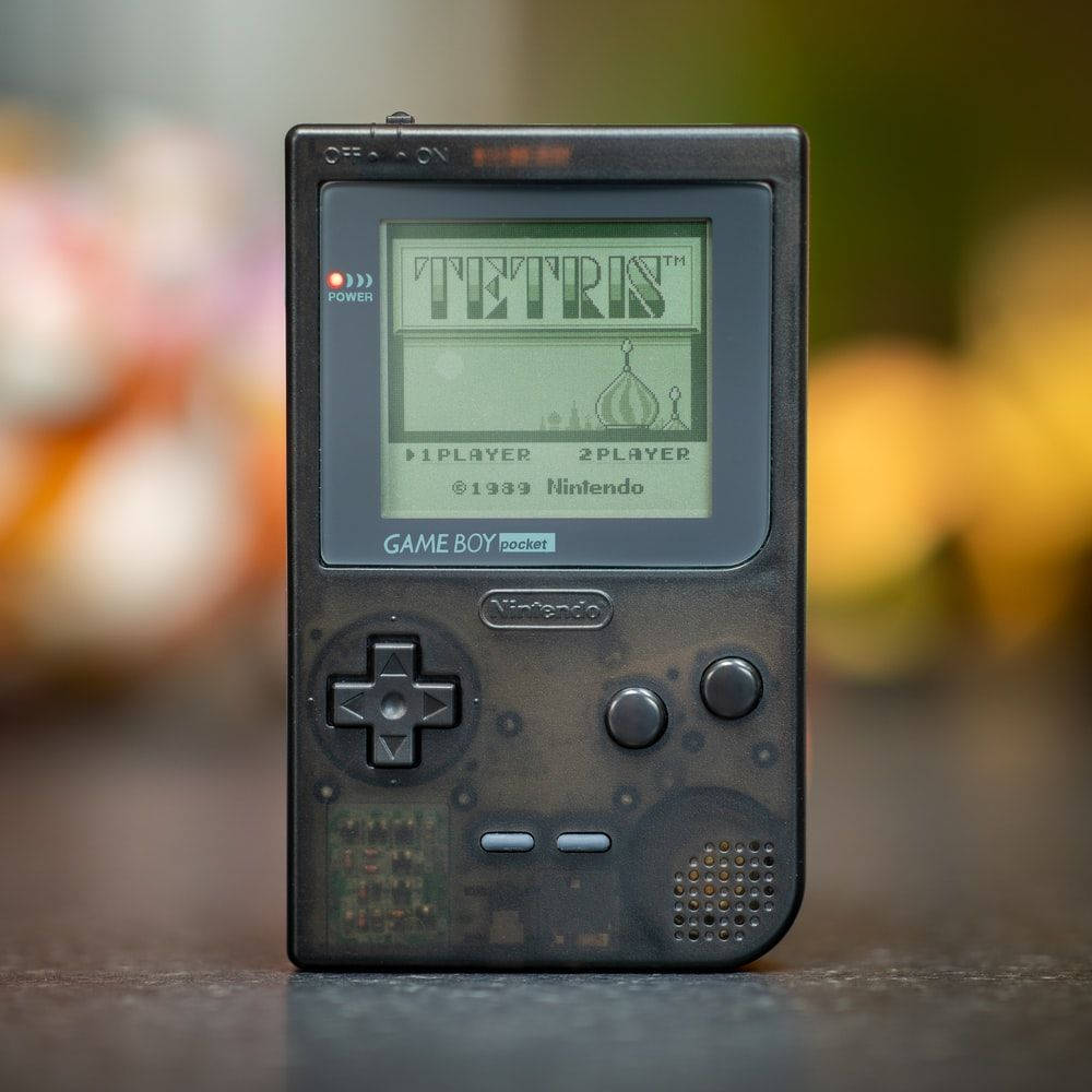 黑色Game Boy Pocket玩《俄羅斯方塊》 壁紙 — 一部復古 Game Boy Pocket 在模糊彩色背景上顯示經典俄羅斯方塊遊戲。