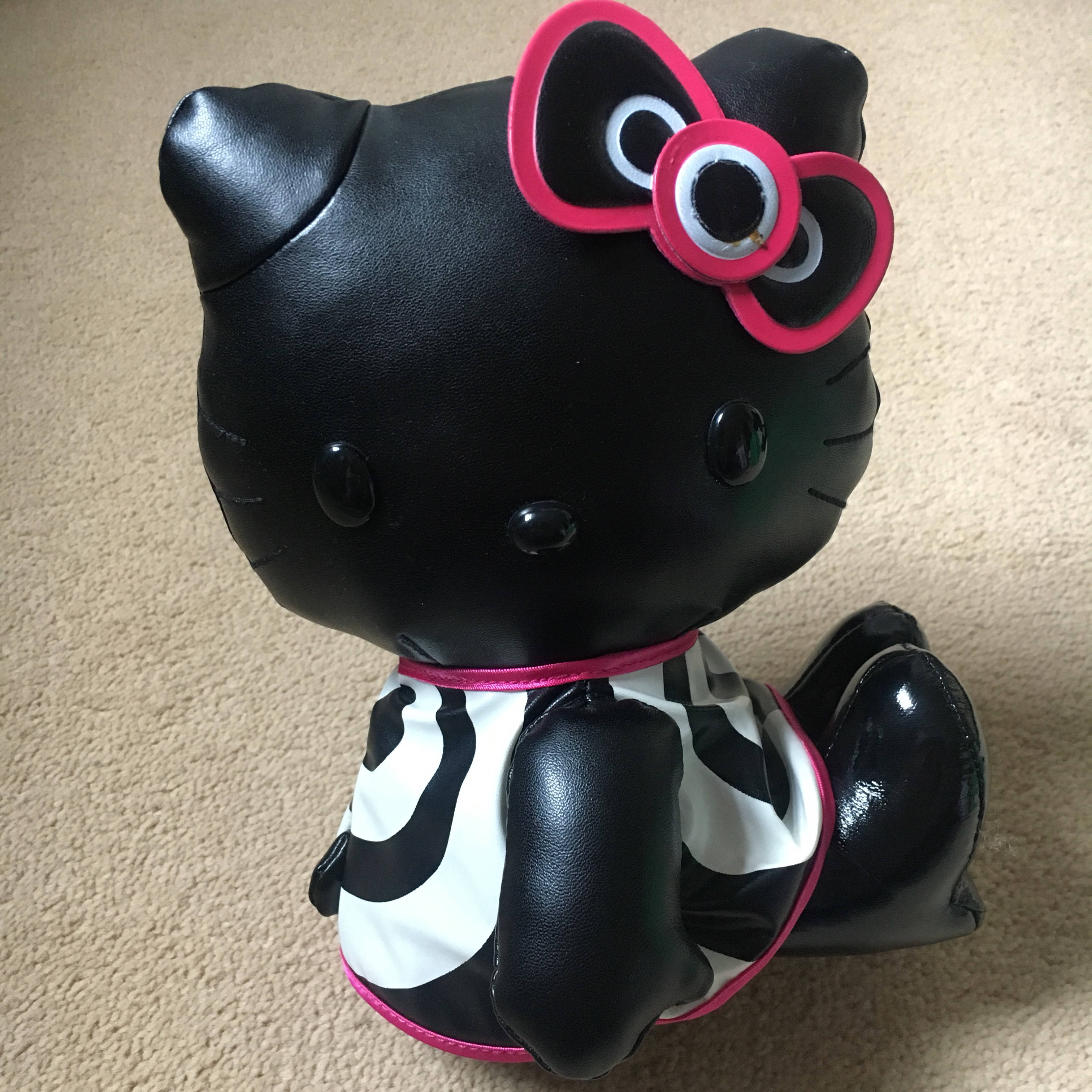黑色Hello Kitty萬聖節玩具 壁紙 — 一隻亮面黑色Hello Kitty毛公仔，戴著鮮粉紅色蝴蝶結，身穿黑白乳牛紋套裝，坐在米色地毯上。