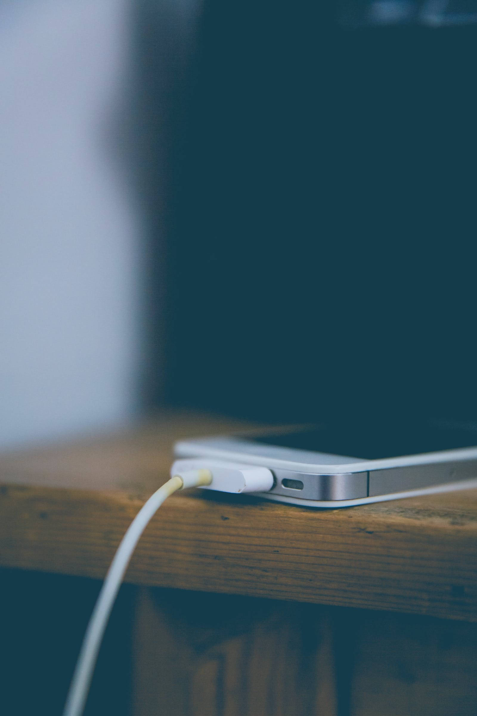 黑色iPhone 4正在充電 壁紙 — 一條白色手機充電線插入一部銀色智能手機，手機放在質樸的木質表面上。