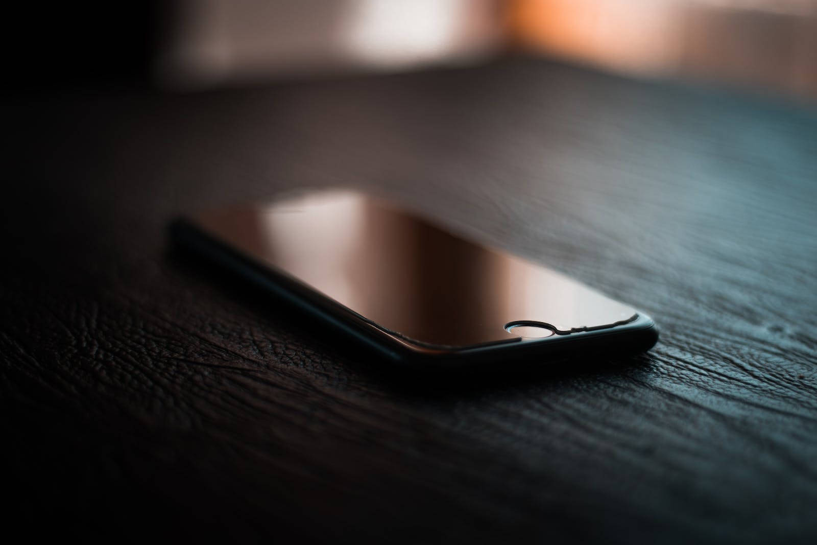 黑色iPhone 7置於桌面 壁紙 — 一部時尚的黑色智能手機放置在深色紋理木質表面上，背景帶有柔和溫暖的燈光。