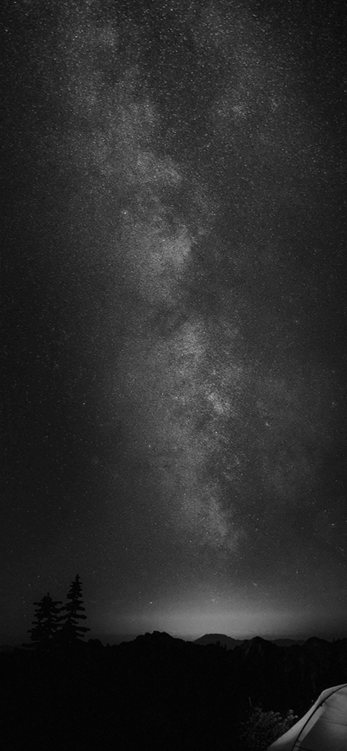 黑色iPhone上的星空夜景 壁紙 — 令人屏息的黑白夜空，佈滿密集的星星，銀河在黑暗的山巒輪廓和帳篷上方拱起。