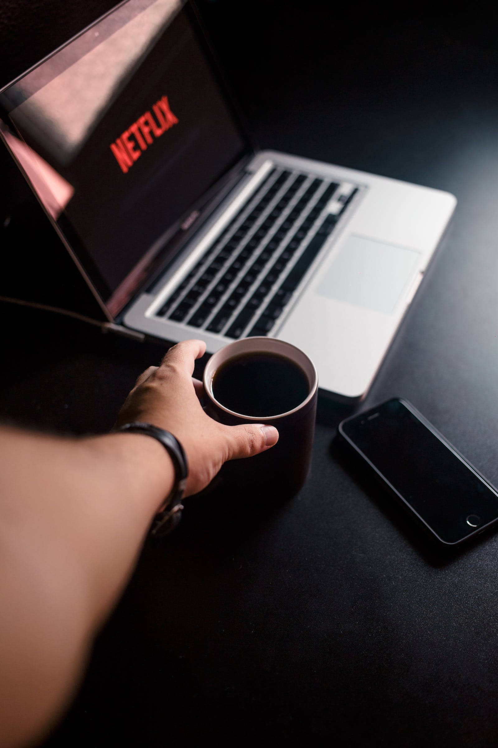 黑色iPhone搭配MacBook與咖啡杯 壁紙 — 一隻手拿著咖啡杯，旁邊是一部顯示Netflix的筆記型電腦，放在深色桌子上，附近有一部手機。