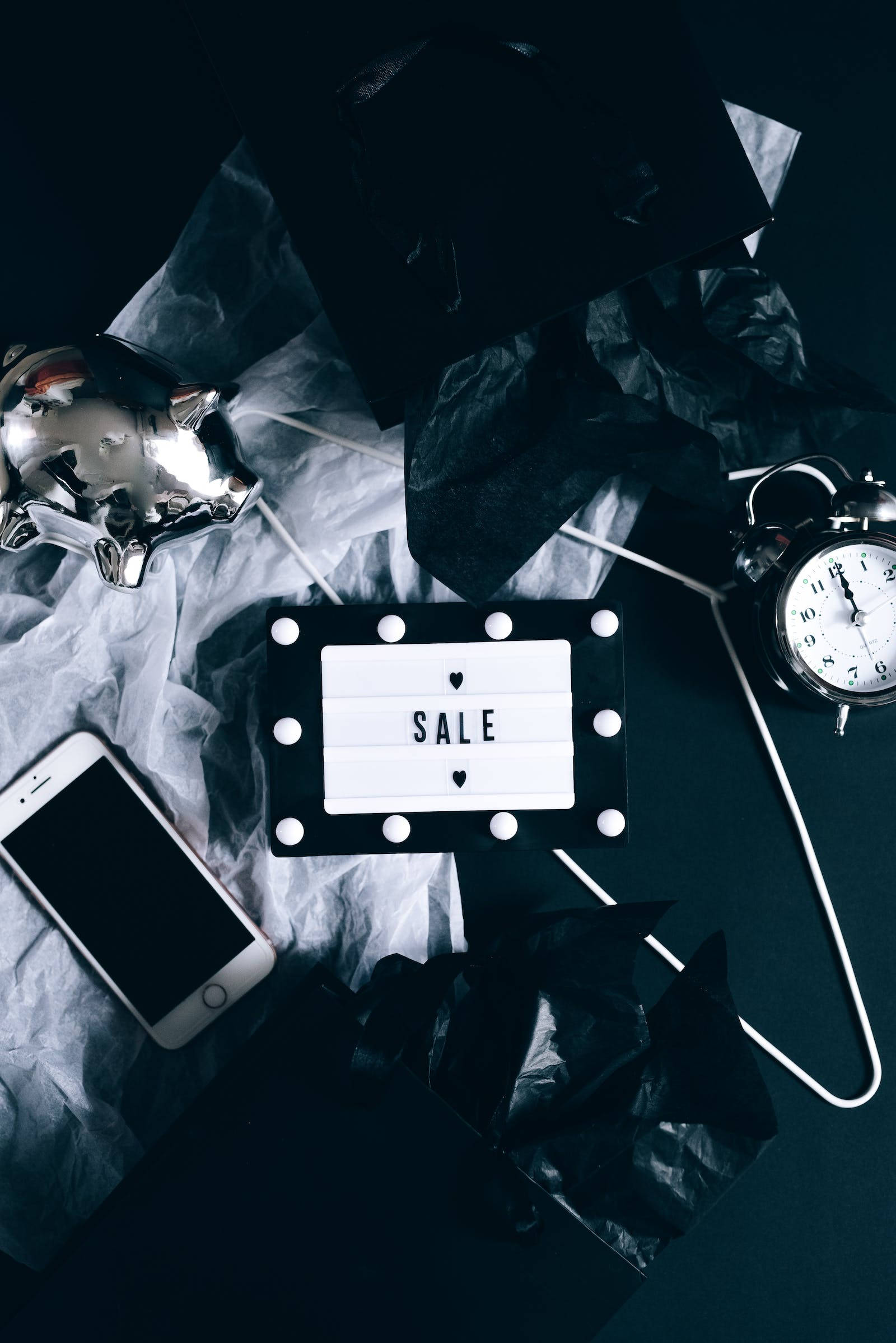 黑色iPhone與其他物品 壁紙 — 一個極簡風格的平鋪構圖，發光的「SALE」標誌周圍環繞著皺巴巴的黑白紙張、一個復古鬧鐘和一部智能手機，置於深色表面上。
