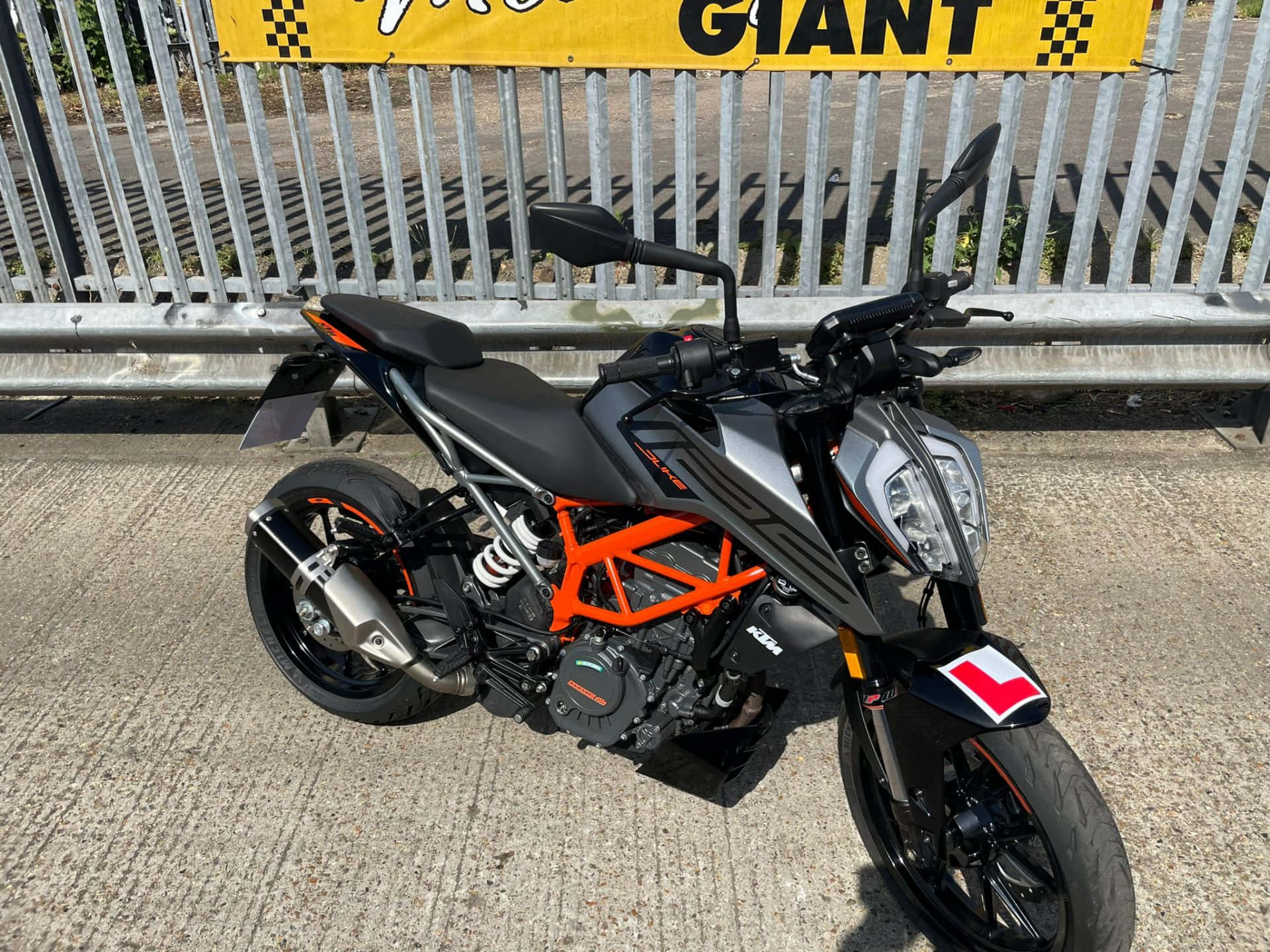 黑色KTM Duke 125停靠金屬大門前 壁紙 — 一架時尚的黑色和橙色KTM電單車在明亮的日光下停泊在金屬圍欄旁邊的混凝土上。