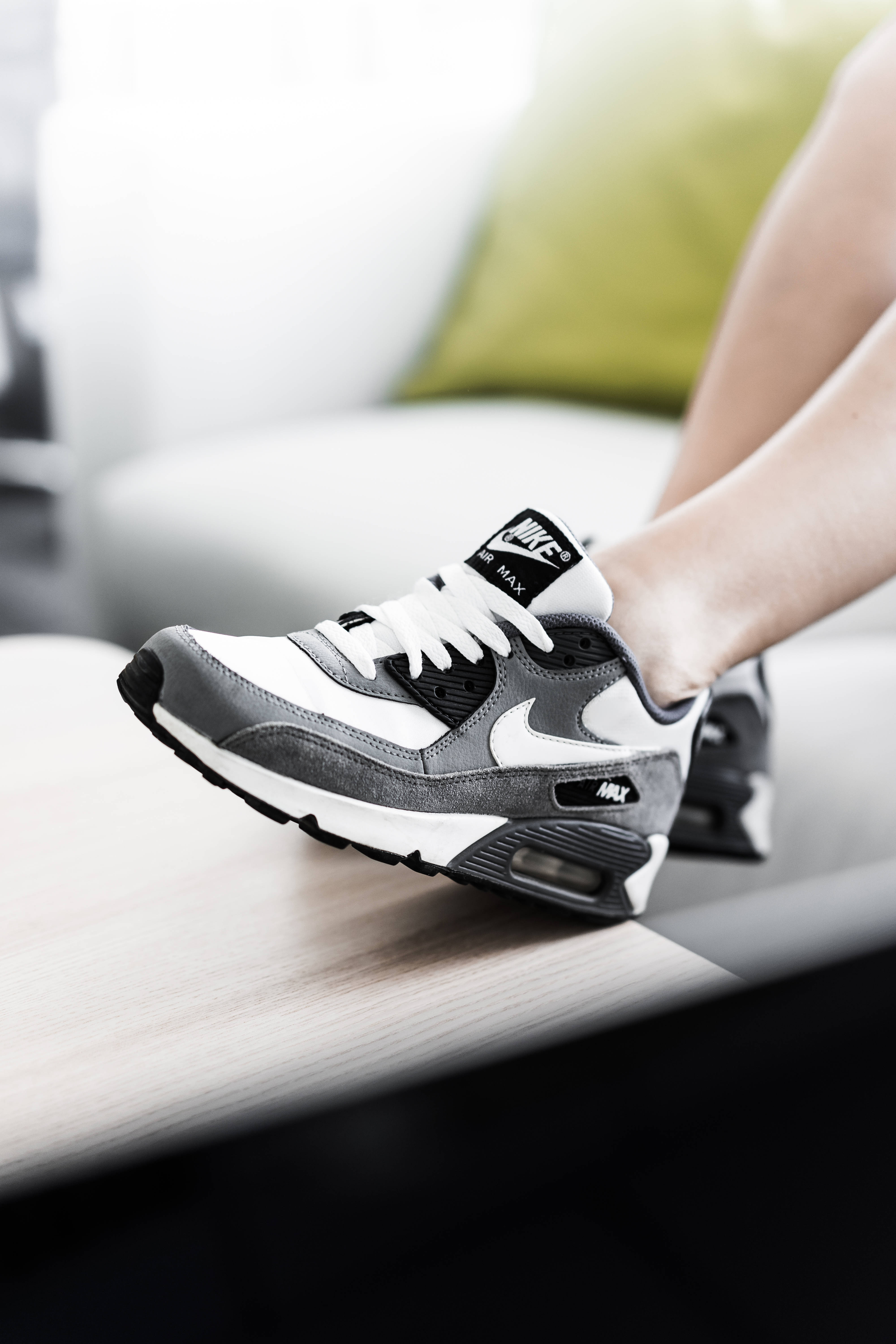 黑色Nike Max鞋 壁紙 — 一個人腳部穿著灰色和白色Nike Air Max運動鞋的特寫，鞋放在木質表面上，背景是模糊的綠色軟墊。