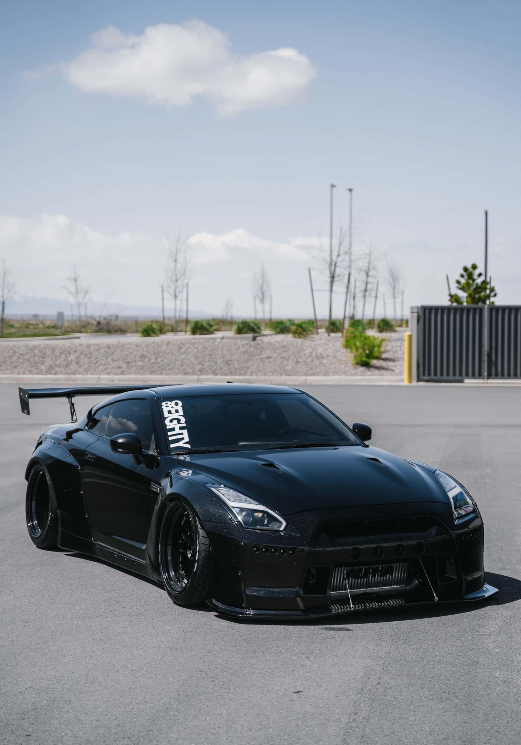 黑色Nissan R35 GT-R停車場圖 壁紙 — 一架型格黑色日產GT-R跑車，配備大型尾翼，停泊在瀝青停車場上，天空局部多雲。