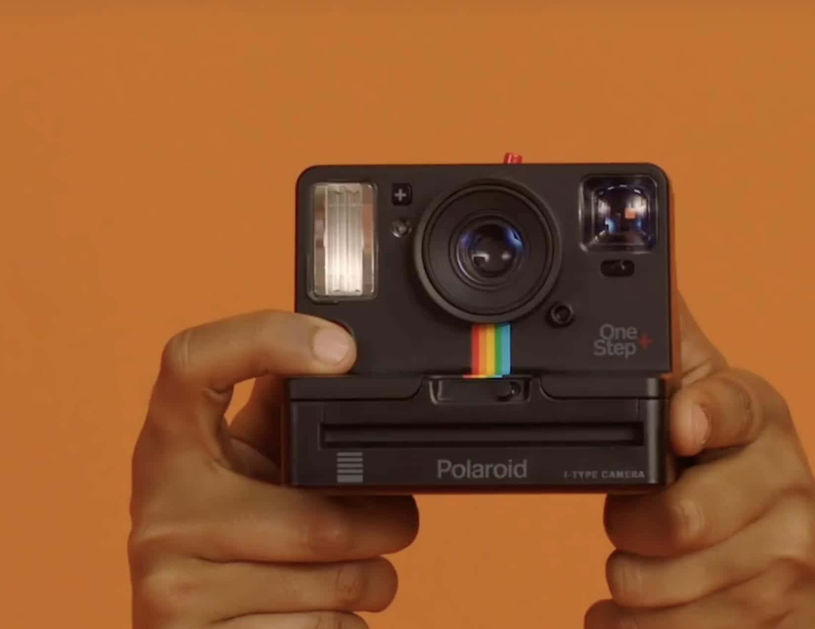雙手拿著一部黑色Polaroid One Step+即影即有相機，背景為橙色。