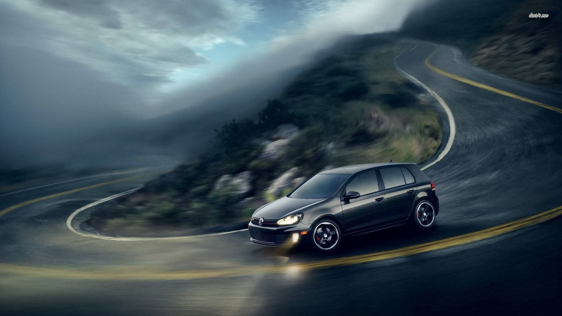 黑色Volkswagen Golf GTI Compact 壁紙 — 一架時尚的深灰色汽車在陰沉多雲的天空下，沿著蜿蜒的山路高速行駛，帶有動態模糊效果。