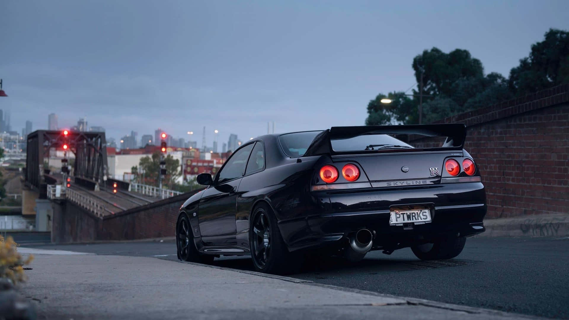 黑色低調R33 GTR 壁紙 — 一架時尚的黑色日產Skyline GT-R在黃昏時停泊在橋上，帶有發光的紅色尾燈和城市天際線背景。
