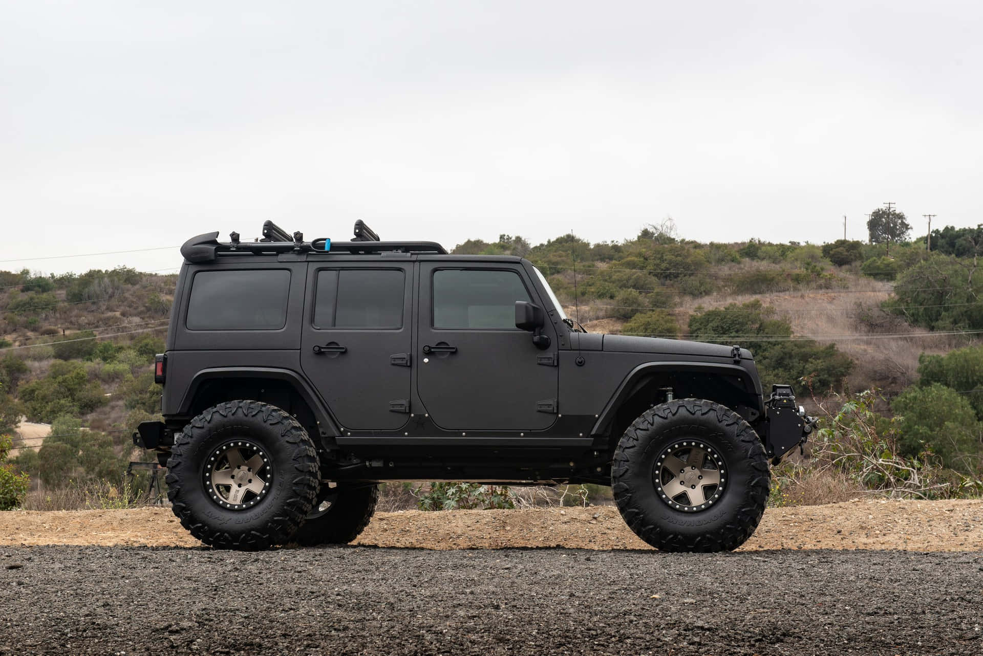 啞光黑色升高Jeep Wrangler停泊在碎石上，背景有灌木山丘。