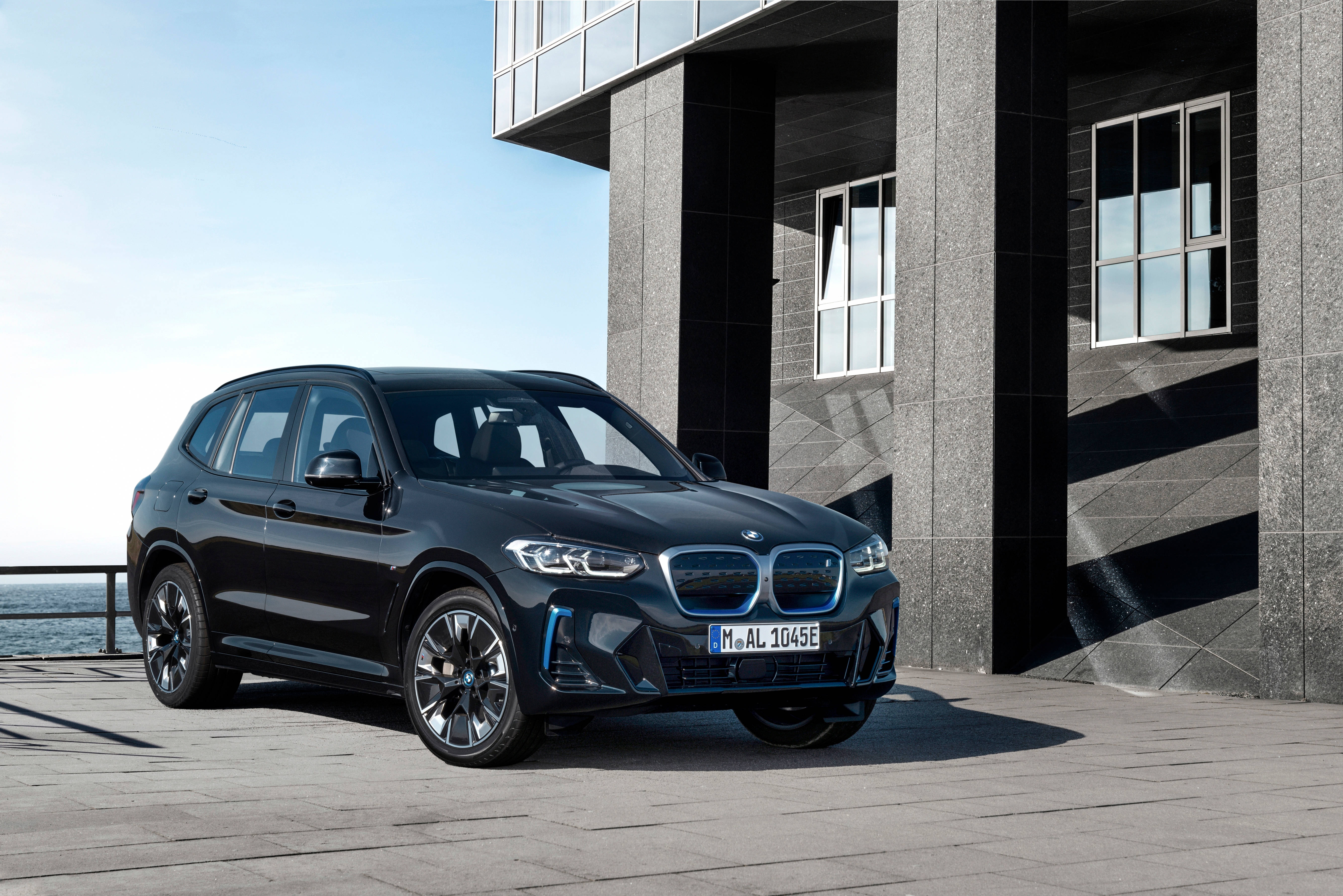 黑色寶馬 iX3 M Sport 壁紙 — 一輛時尚的黑色BMW電動SUV停在一棟現代混凝土建築旁，享有晴朗藍天下的海景。