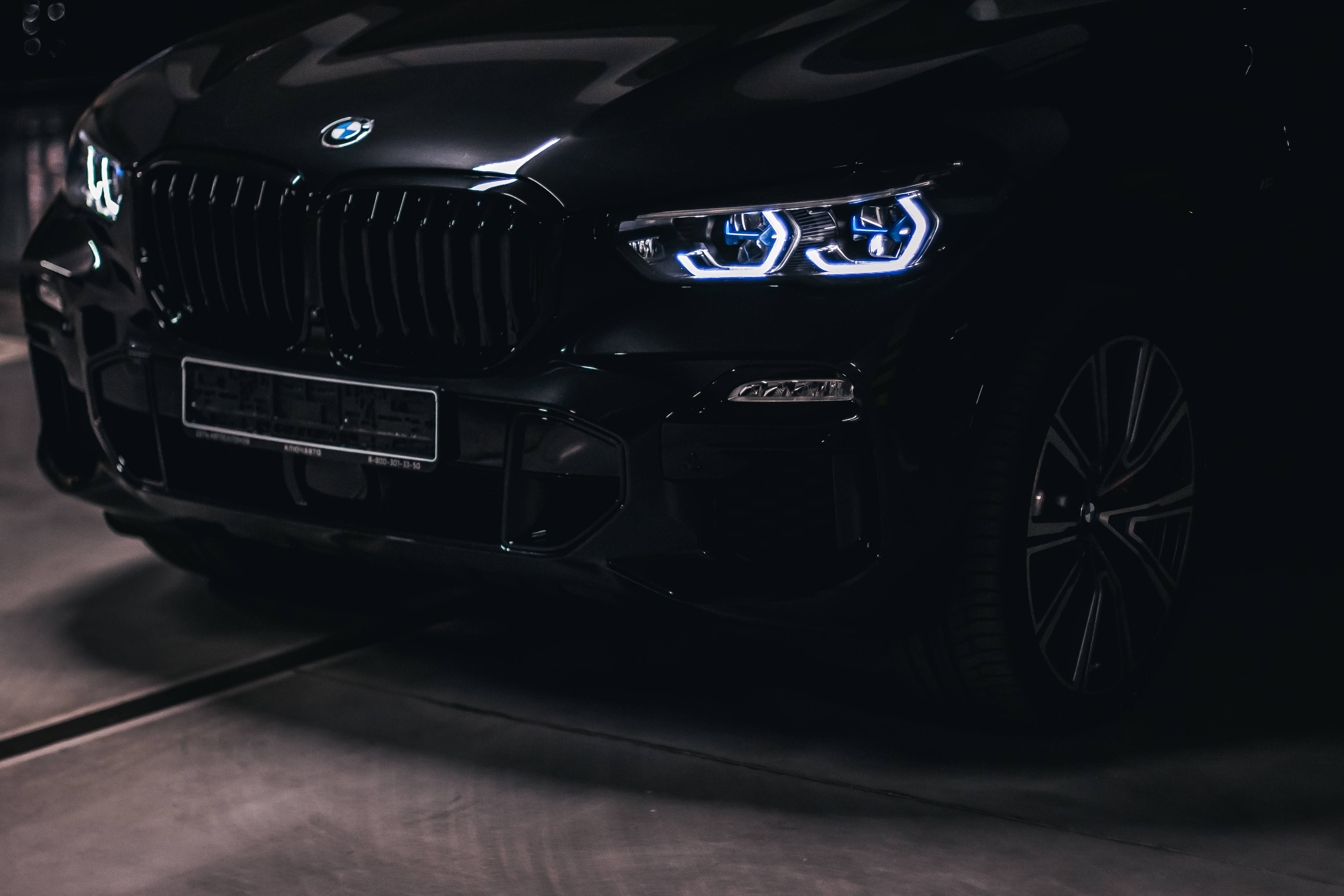 黑色寶馬車頭燈與保險桿 壁紙 — 一輛時尚的黑色BMW SUV在昏暗的車庫中發出銳利的藍色車頭燈光，在混凝土上投射出戲劇性的陰影。
