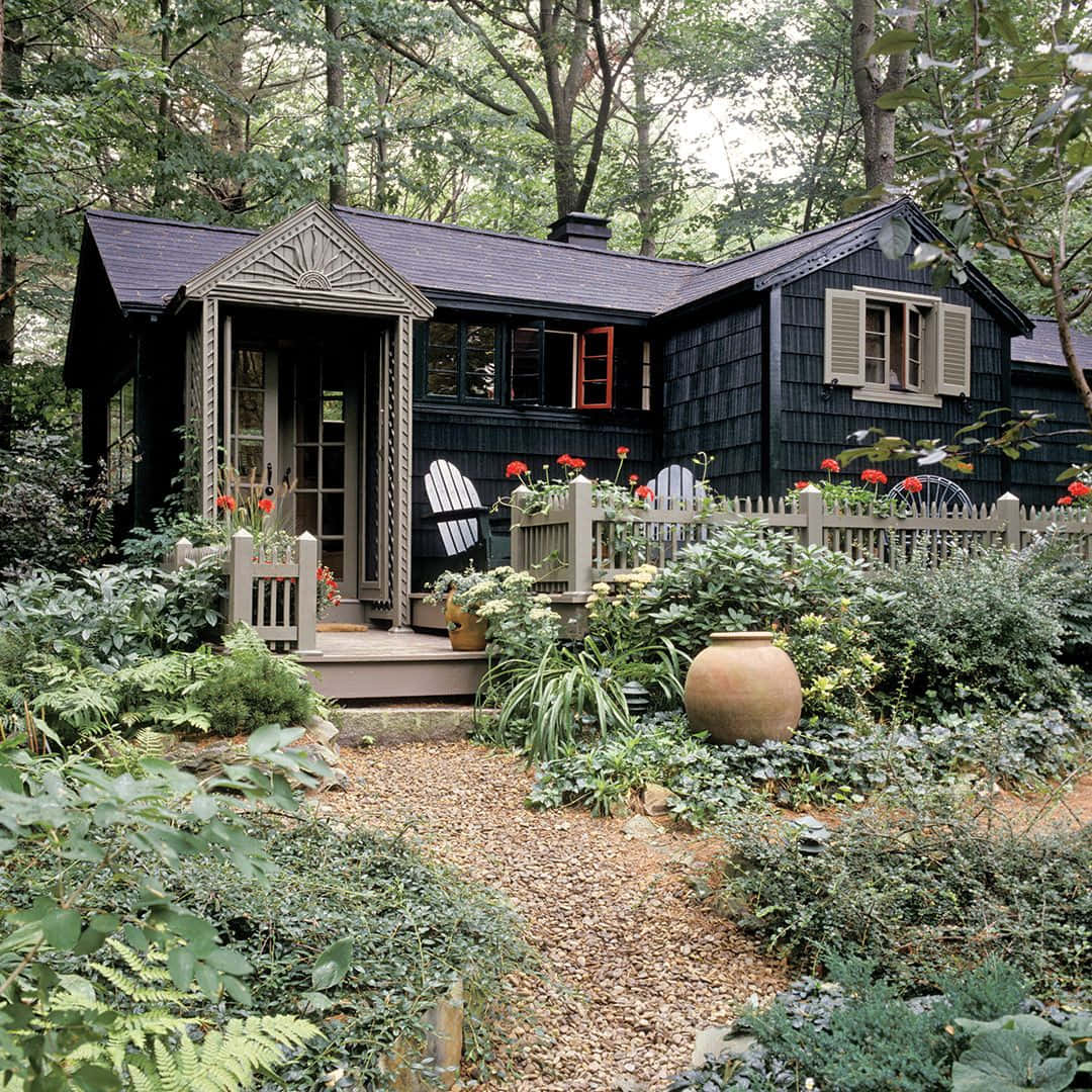 黑色小屋攝影圖 壁紙 — 一間迷人的黑色木瓦小屋，坐落在鬱鬱蔥蔥的綠色森林中，一條碎石路通向點綴著紅花和白椅的門廊。