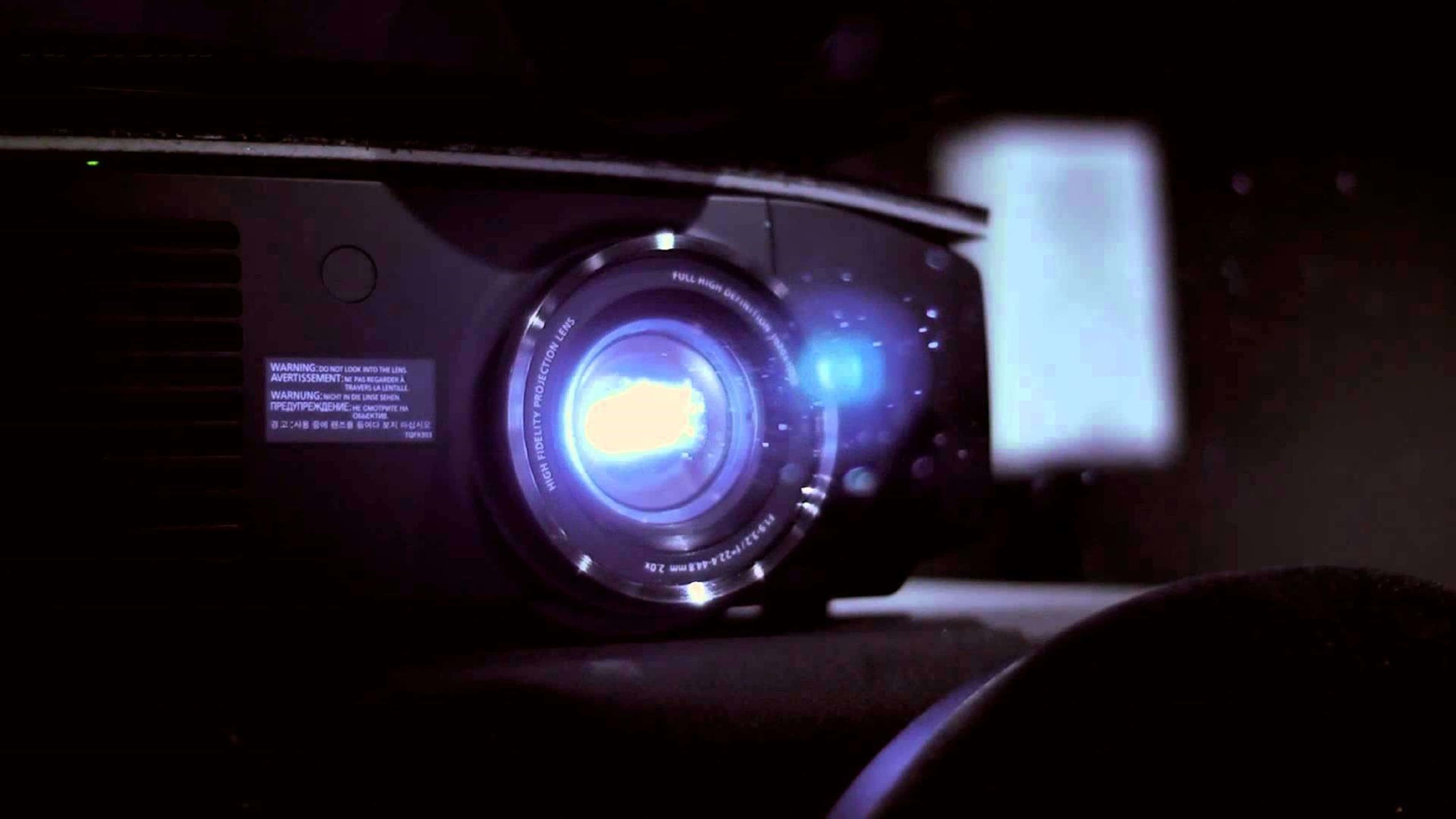4K Projector 桌布 壁紙 — 黑色投影機在黑暗房間中發出藍光，在屏幕上投射出微弱圖像的特寫。
