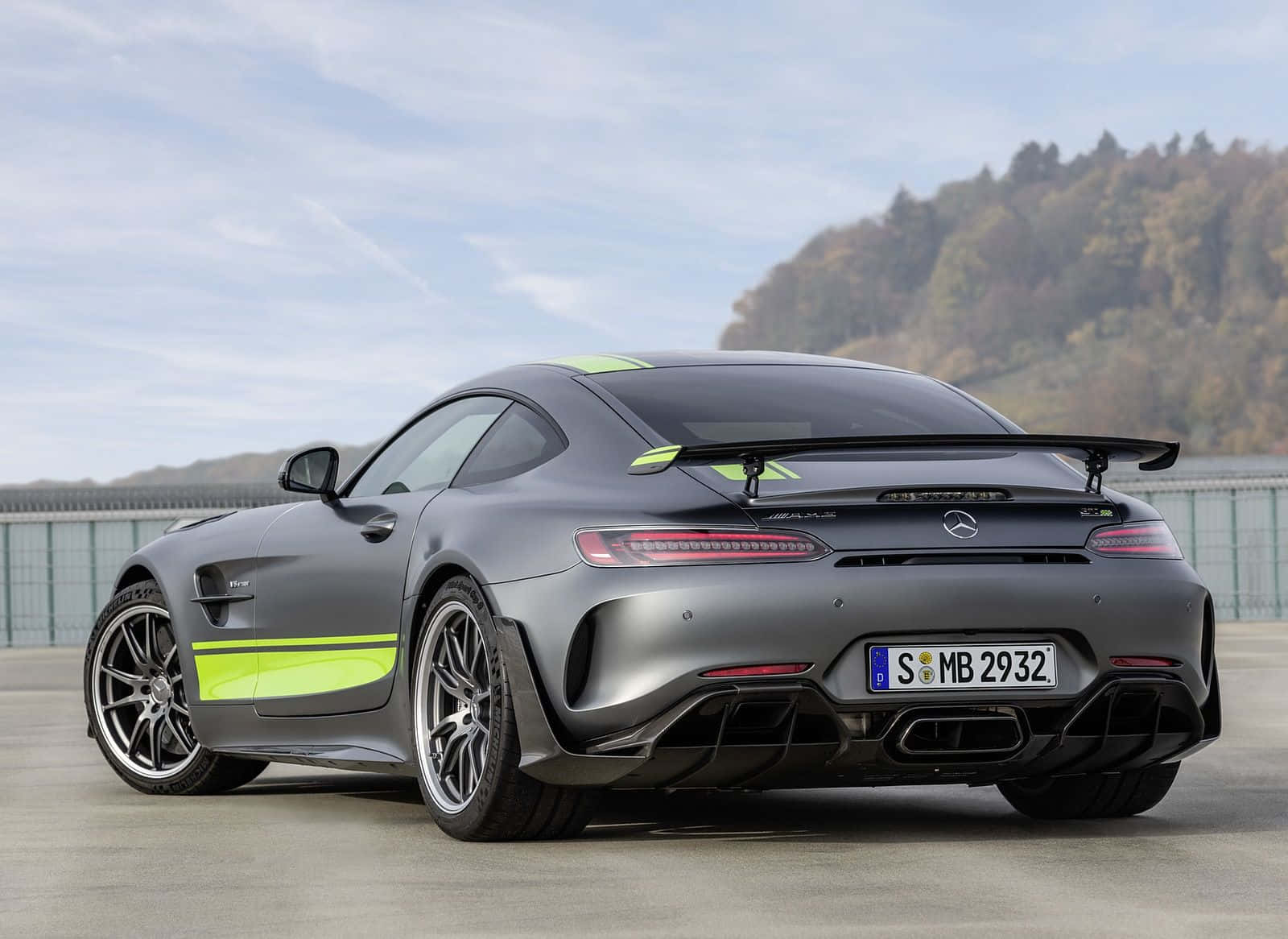 黑色梅賽德斯-奔馳AMG GT R Pro罕見角度背景 壁紙 — 一輛啞光灰色的梅賽德斯-AMG GT R 跑車，帶有螢光綠色點綴，停放在混凝土表面上，背景是森林覆蓋的山丘，天空部分多雲。
