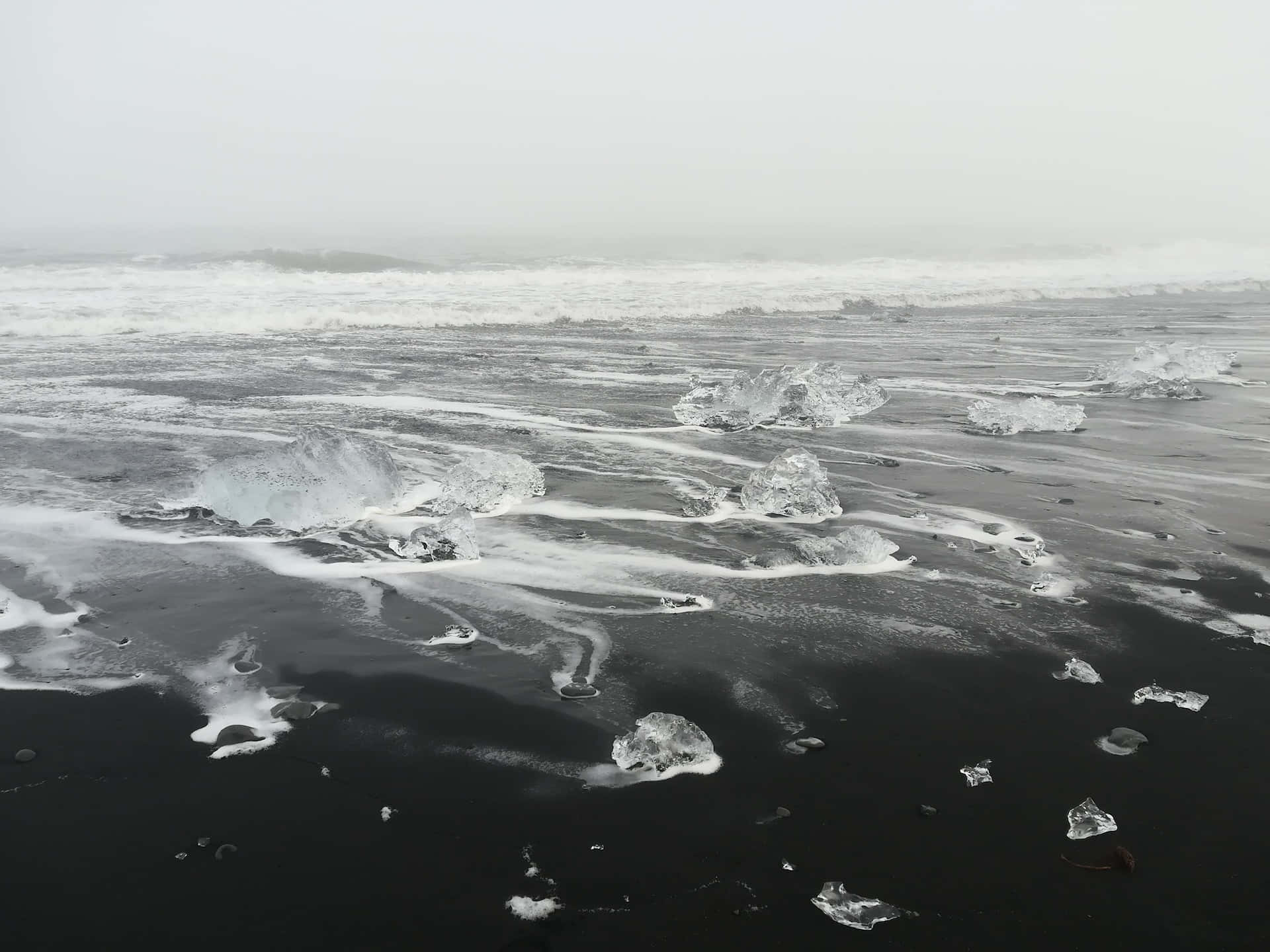 Black And White Beach 桌布 壁紙 — 黑色火山沙灘上散落著半透明的冰塊，海浪在陰天中拍打。