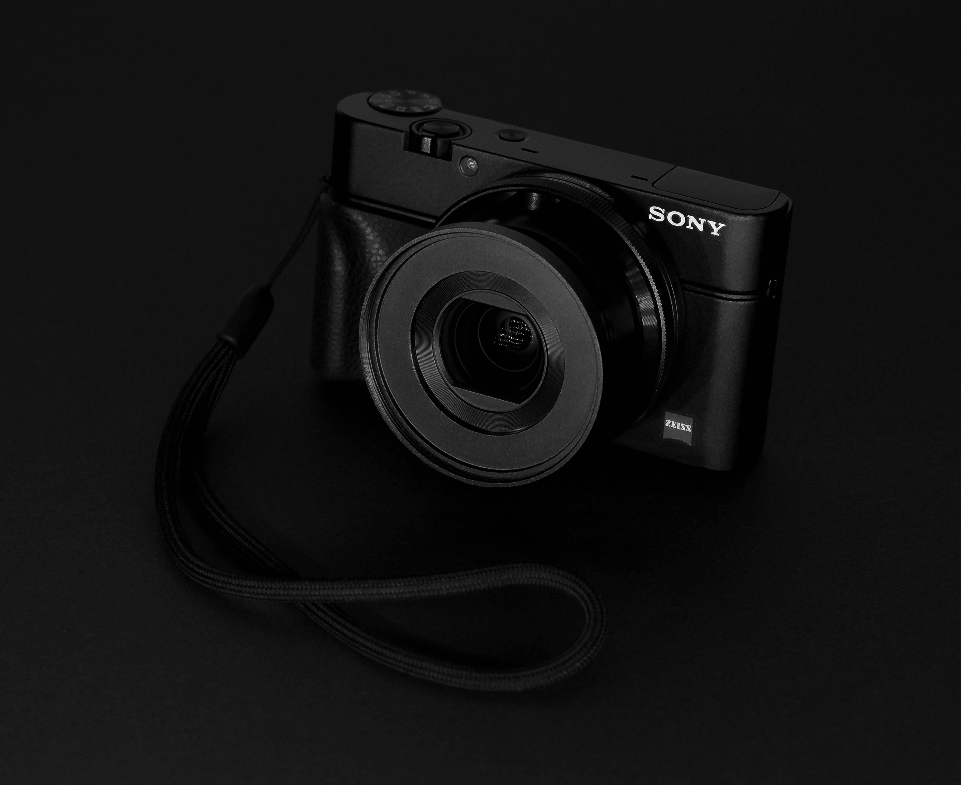 黑色索尼數碼相機 壁紙 — 一部時尚的黑色Sony相機配備Zeiss鏡頭，放置在深色表面上，其背帶纏繞在一旁，在戲劇性的工作室燈光下。