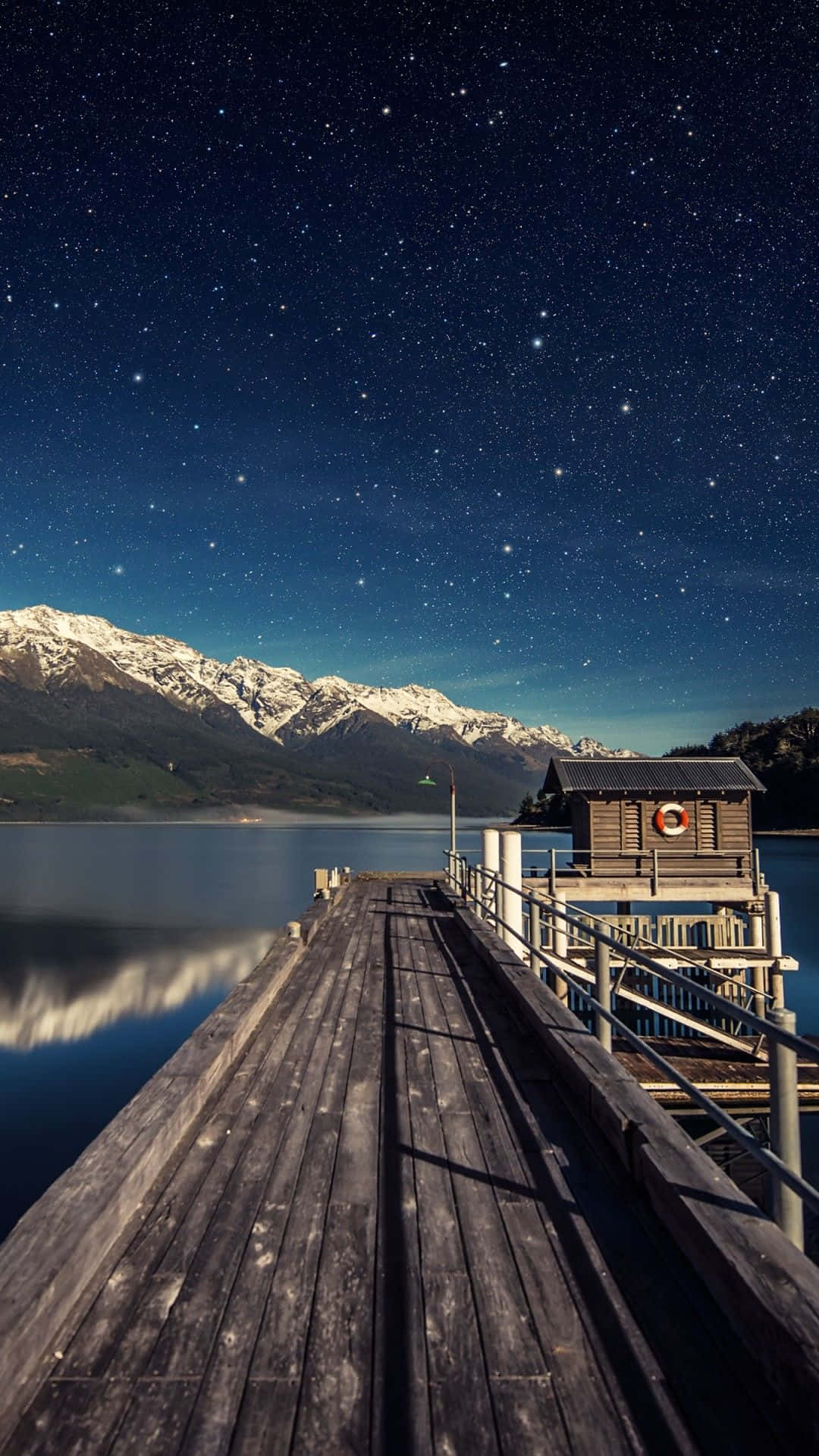 黑色背景上的抽象多彩波浪 壁紙 — 木製碼頭延伸至星空下的夜晚湖泊，平靜的水面倒映著積雪的山峰。