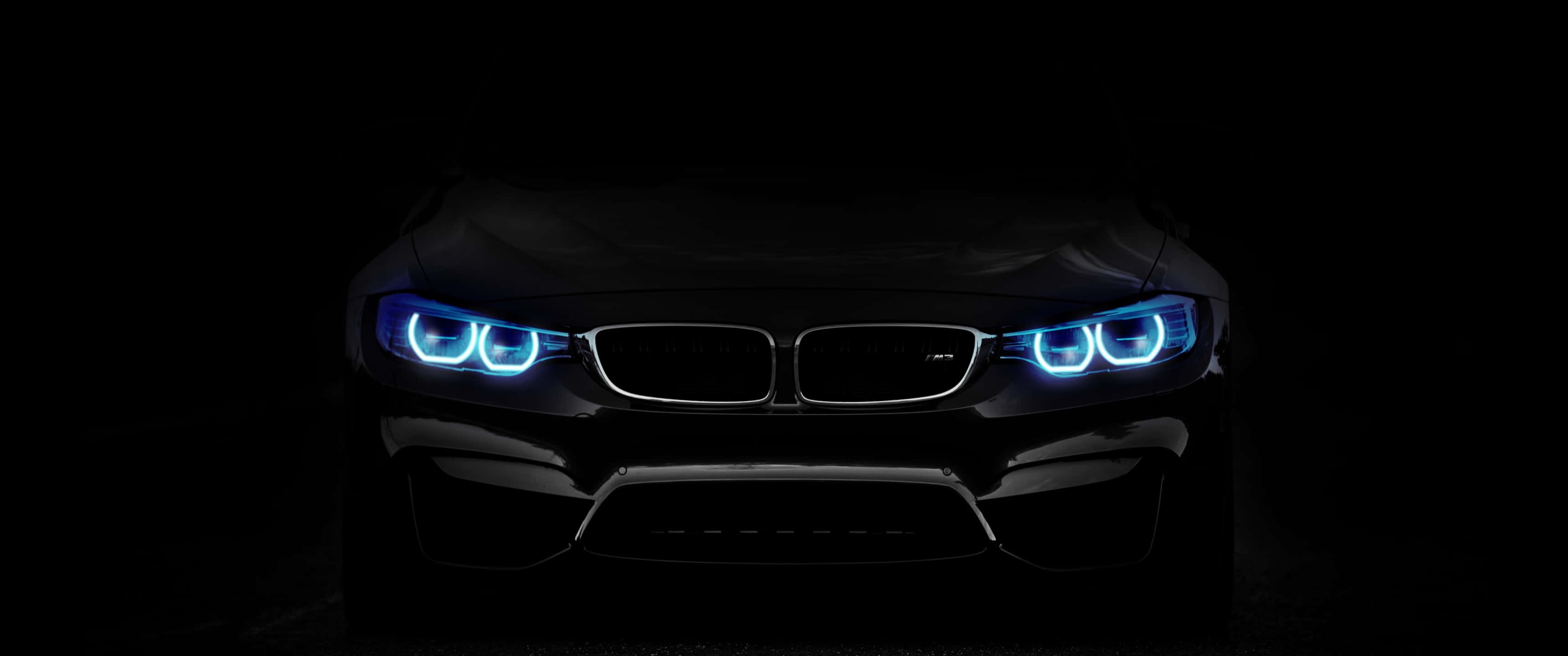 黑色藍色 BMW 3440x1440p 背景 壁紙 — 一輛時尚的黑色BMW跑車在漆黑背景下散發出鮮豔的藍色車頭燈光芒。