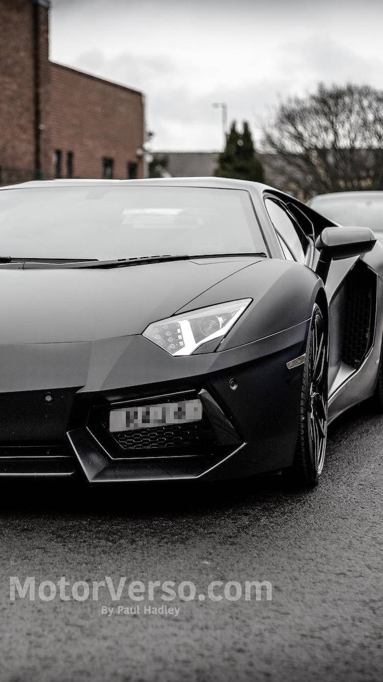 黑色蘭博基尼Aventador汽車iPhone壁紙 壁紙 — 一架時尚的啞光黑色林寶堅尼Aventador停泊在濕漉漉的路面上，背景是磚砌建築和光禿禿的樹木，天空多雲。