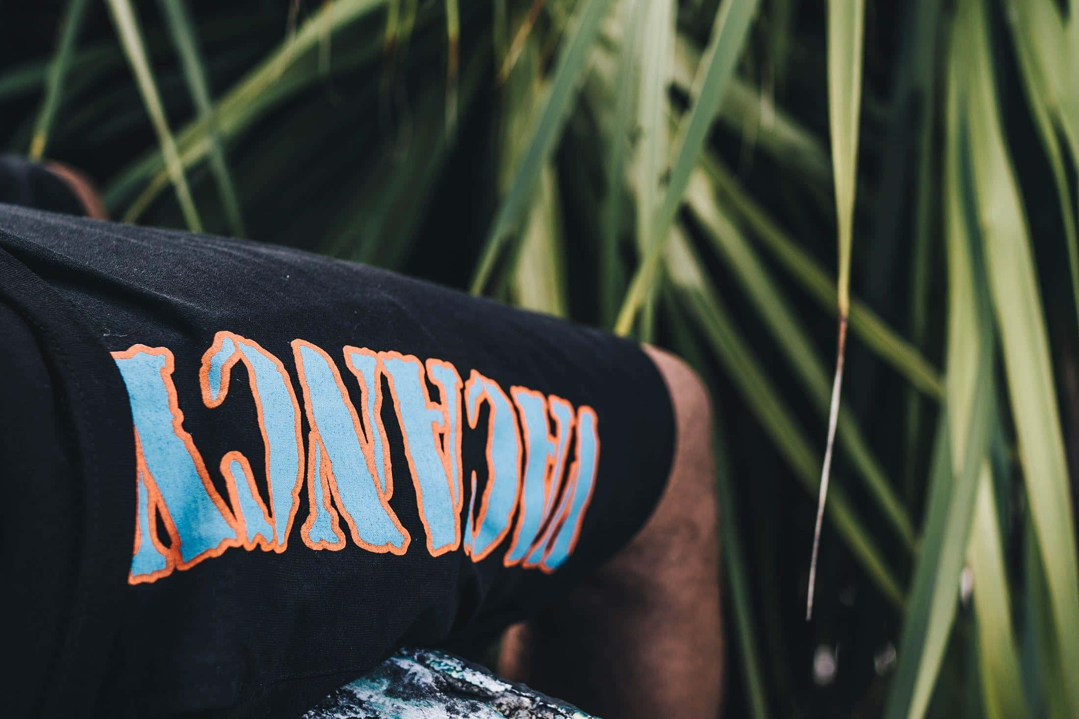 Vlone 桌布 壁紙 — 黑色衣袖特寫，上面有粗體藍色和橙色嘅「VACANCY」文字，背景係模糊嘅綠色樹葉。