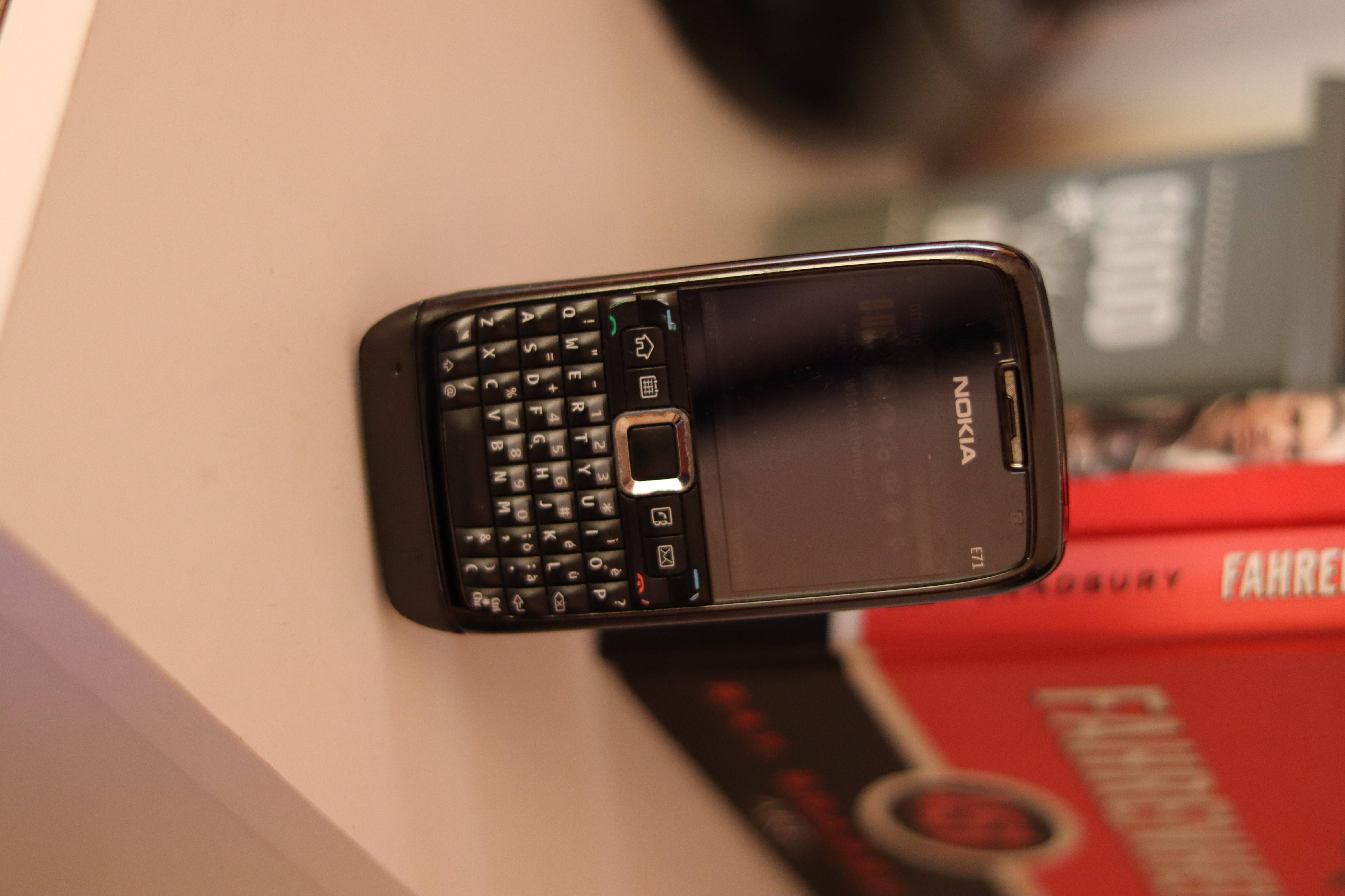 黑色諾基亞 E71 壁紙 — 一部黑色Nokia E71智能手機配備實體鍵盤，放在白色表面上，旁邊有紅色書籍。