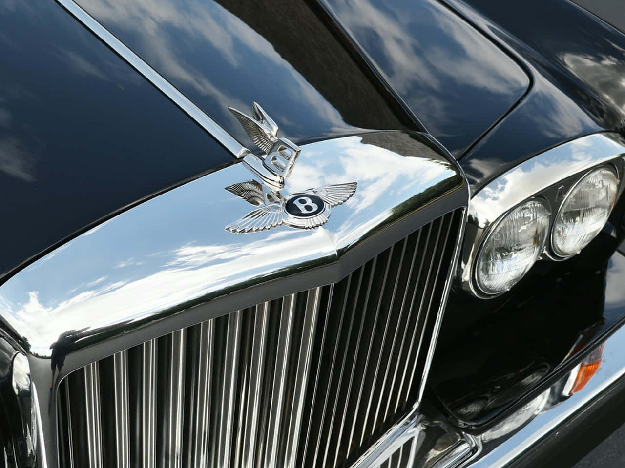 Bentley T2 桌布 壁紙 — 黑色賓利閃亮鍍鉻格柵及帶翼 'B' 徽章的特寫，反射明亮天空。