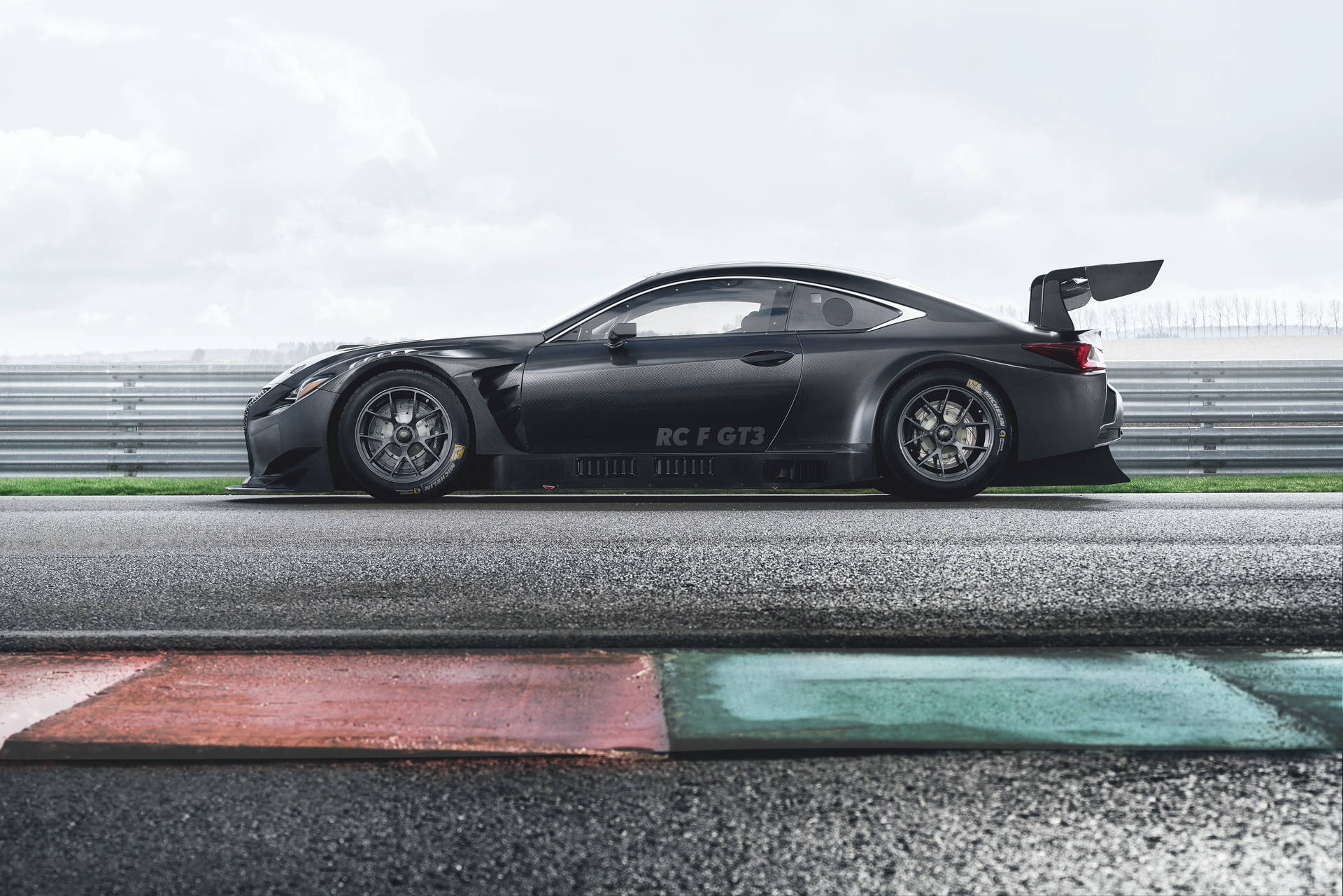 啞光黑色 Lexus RC F GT3 賽車在濕滑賽道上，有紅色和綠色路緣石。