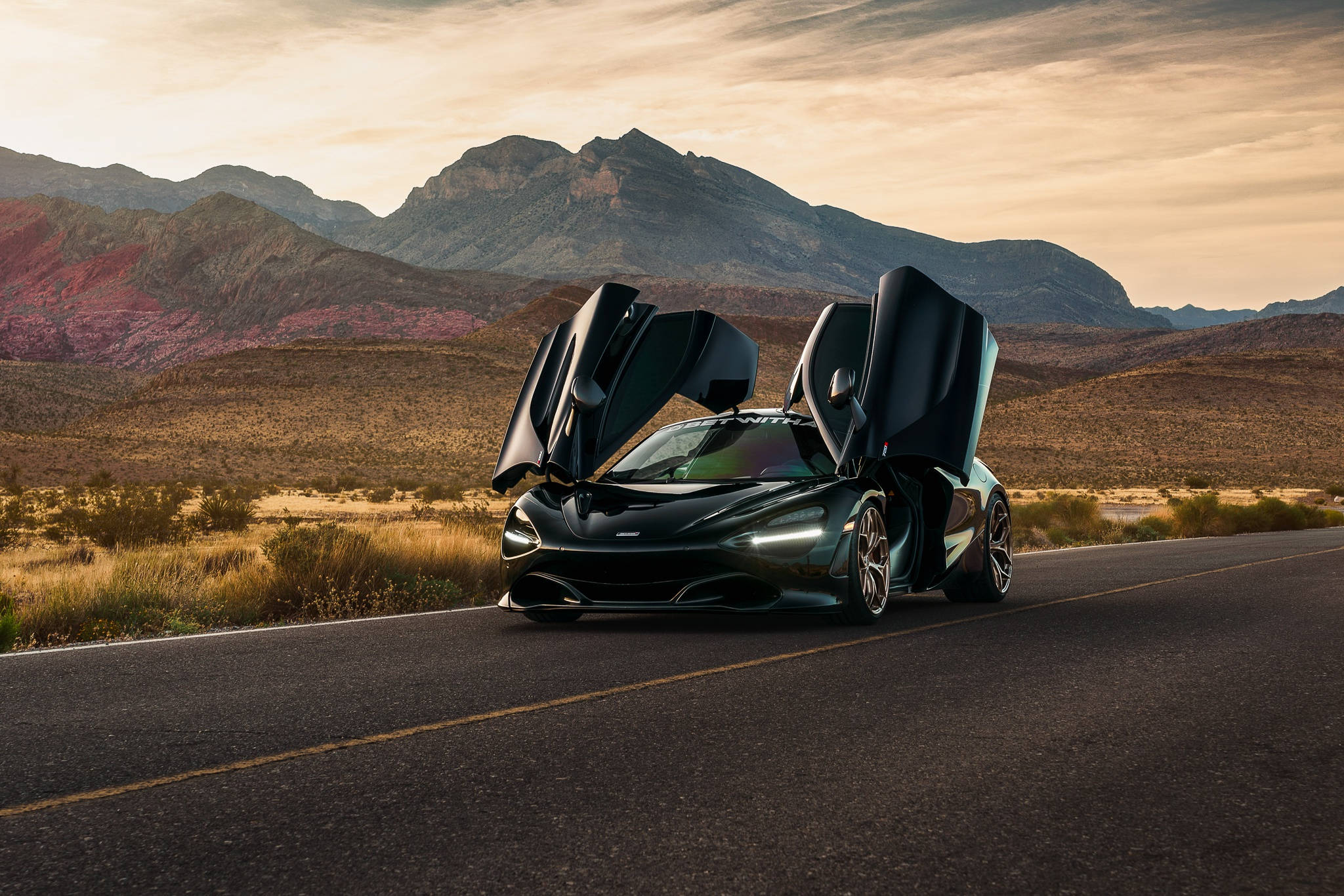 黑色麥拉倫720s停泊於沙漠公路 壁紙 — 一架線條流暢的黑色McLaren跑車，鷗翼車門敞開，停在沙漠公路上，背景是崎嶇的山脈，溫暖的夕陽映照天空。