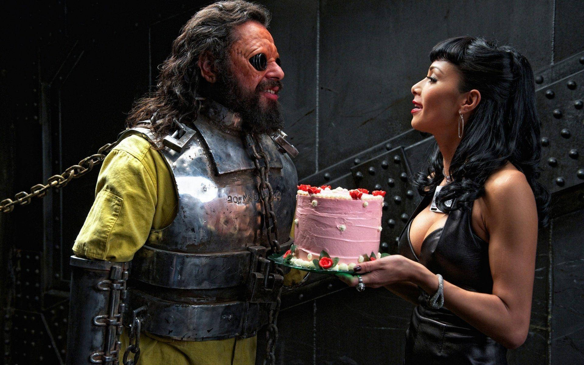 《黑衣人3》反派角色 壁紙 — 一位身穿黑色皮革的女人，在深色金屬牆前，向一位留鬍鬚、身穿生鏽盔甲的男人遞上粉紅色蛋糕。