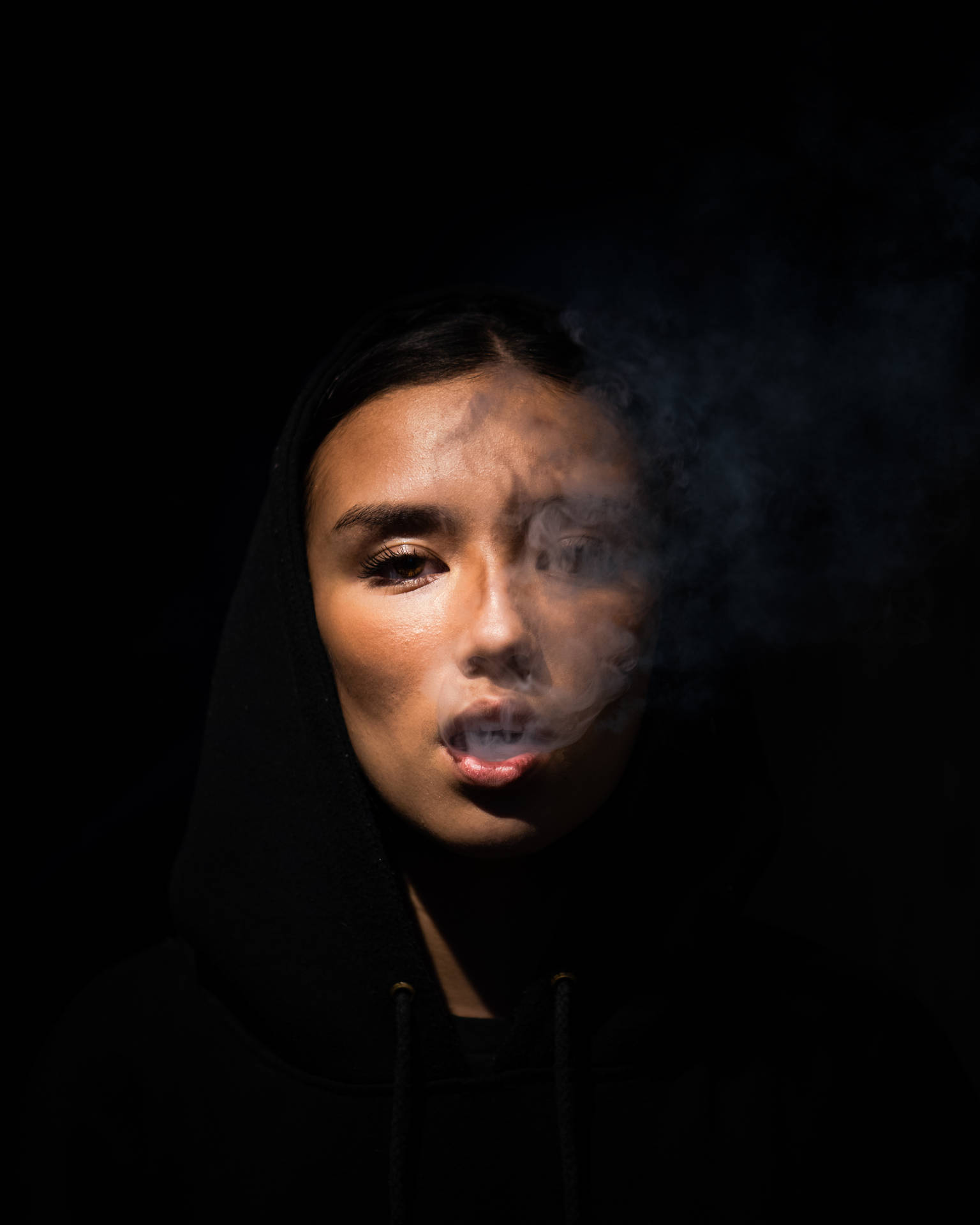 穿著黑色連帽衫的人在黑暗工作室燈光下呼出煙霧。