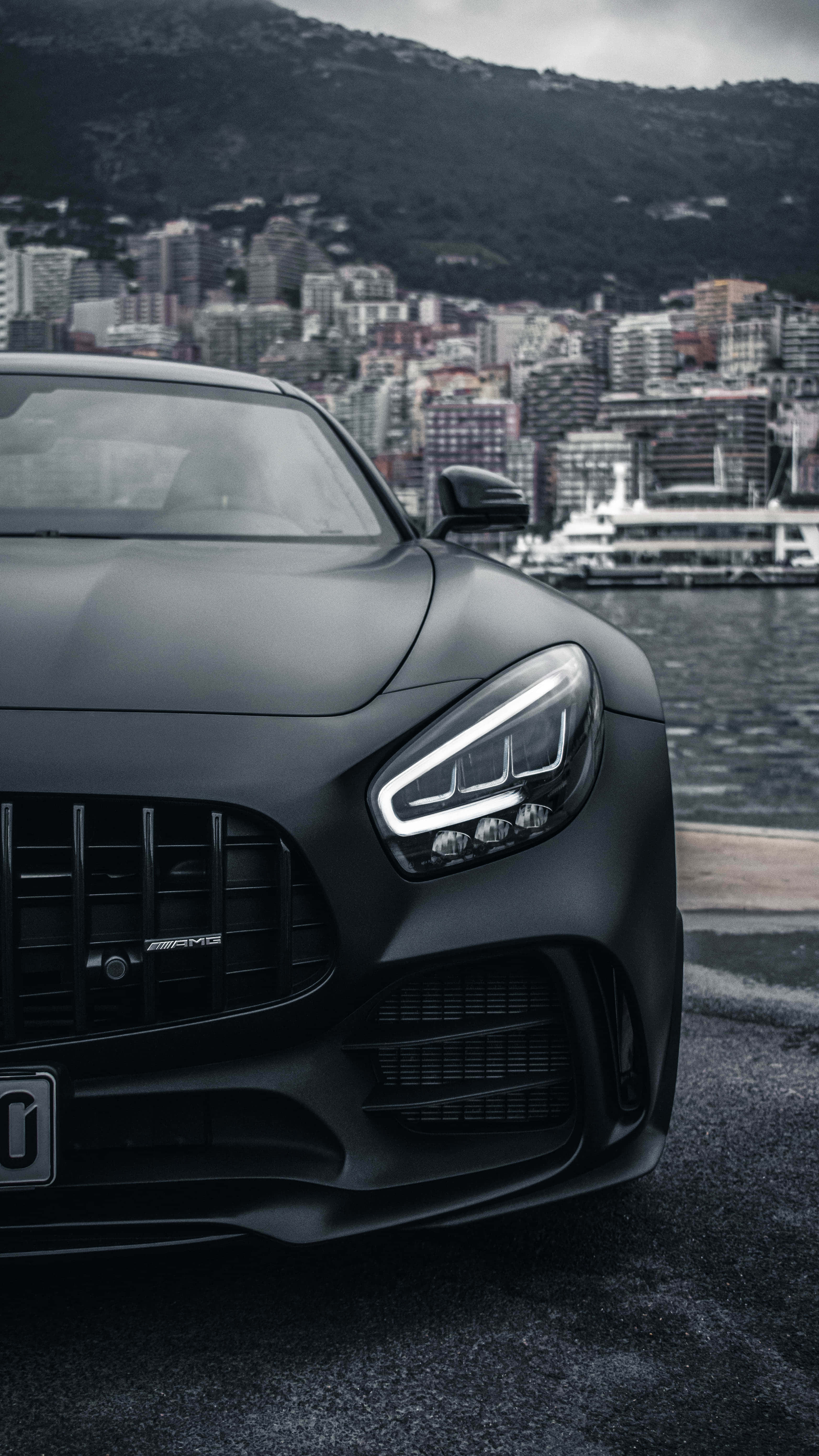黑調裁切安卓AMG GT-R背景 壁紙 — 一架啞光黑色 Mercedes-AMG 跑車停泊在海濱，背景是城市天際線和山脈。