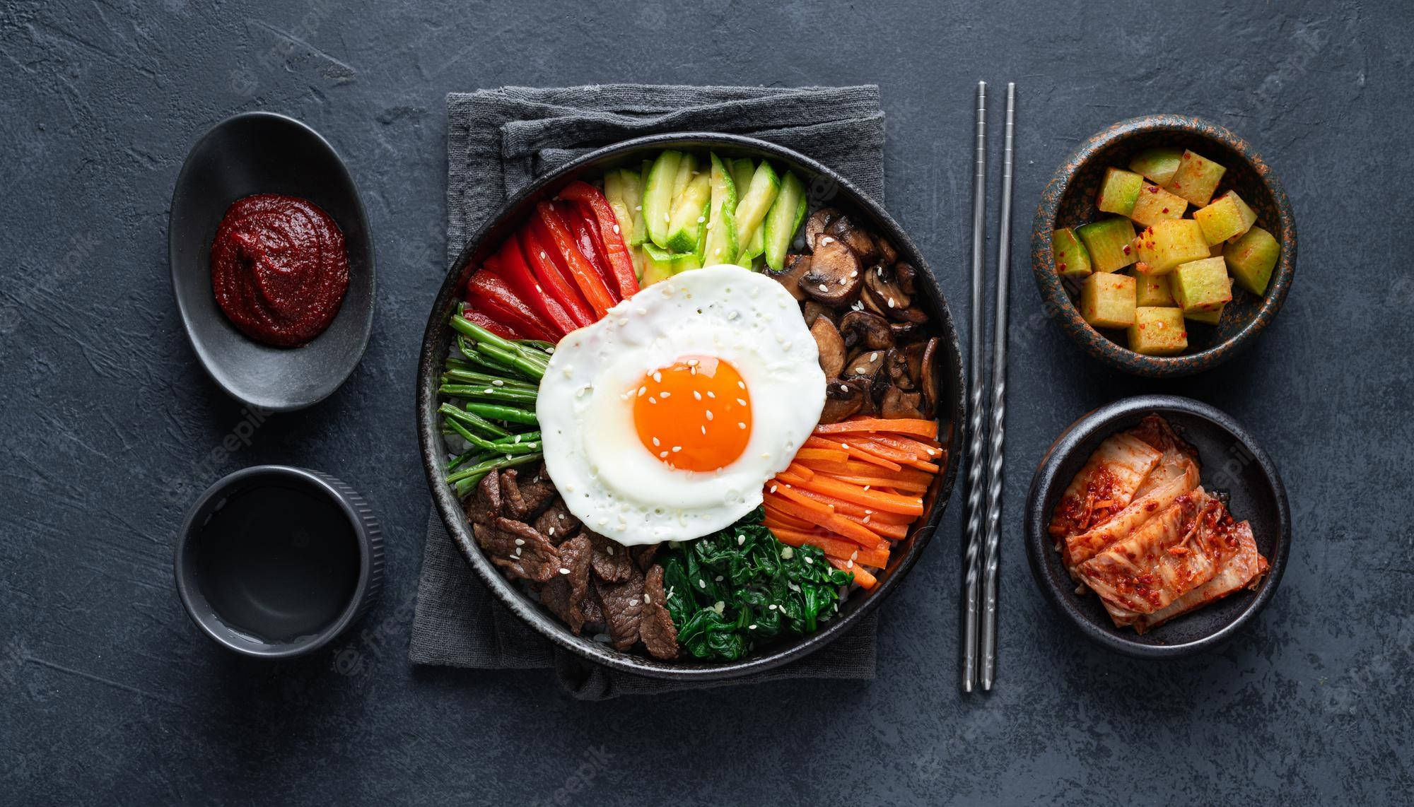黑調韓式拌飯美食圖 壁紙 — 一個色彩繽紛的韓國石鍋拌飯，上面放著太陽蛋和各種蔬菜，置於深色石面上。