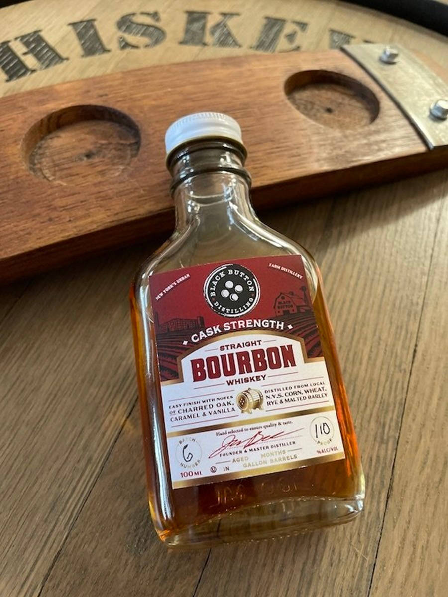 黑鈕釦蒸餾廠桶強波本威士忌 壁紙 — 一小瓶 Black Button Cask Strength 波本威士忌放在木桌上，旁邊有一個威士忌桶杯墊。