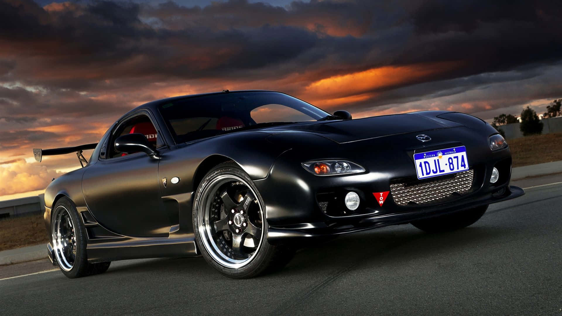 黑色Mazda RX-7跑車在日落時分行駛在道路上，天空戲劇性。