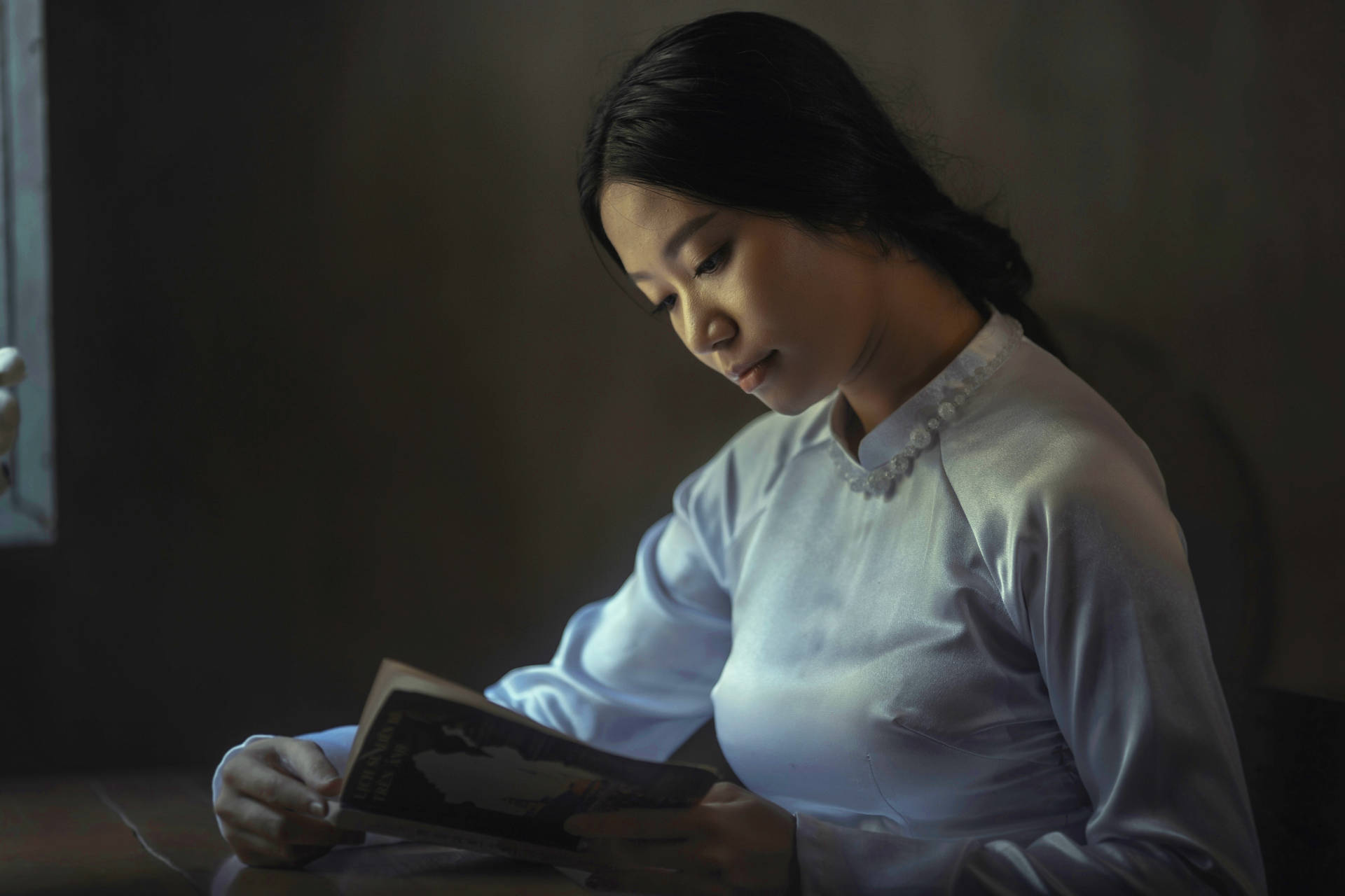 女性在安靜昏暗的房間裡藉著窗光閱讀一本書。